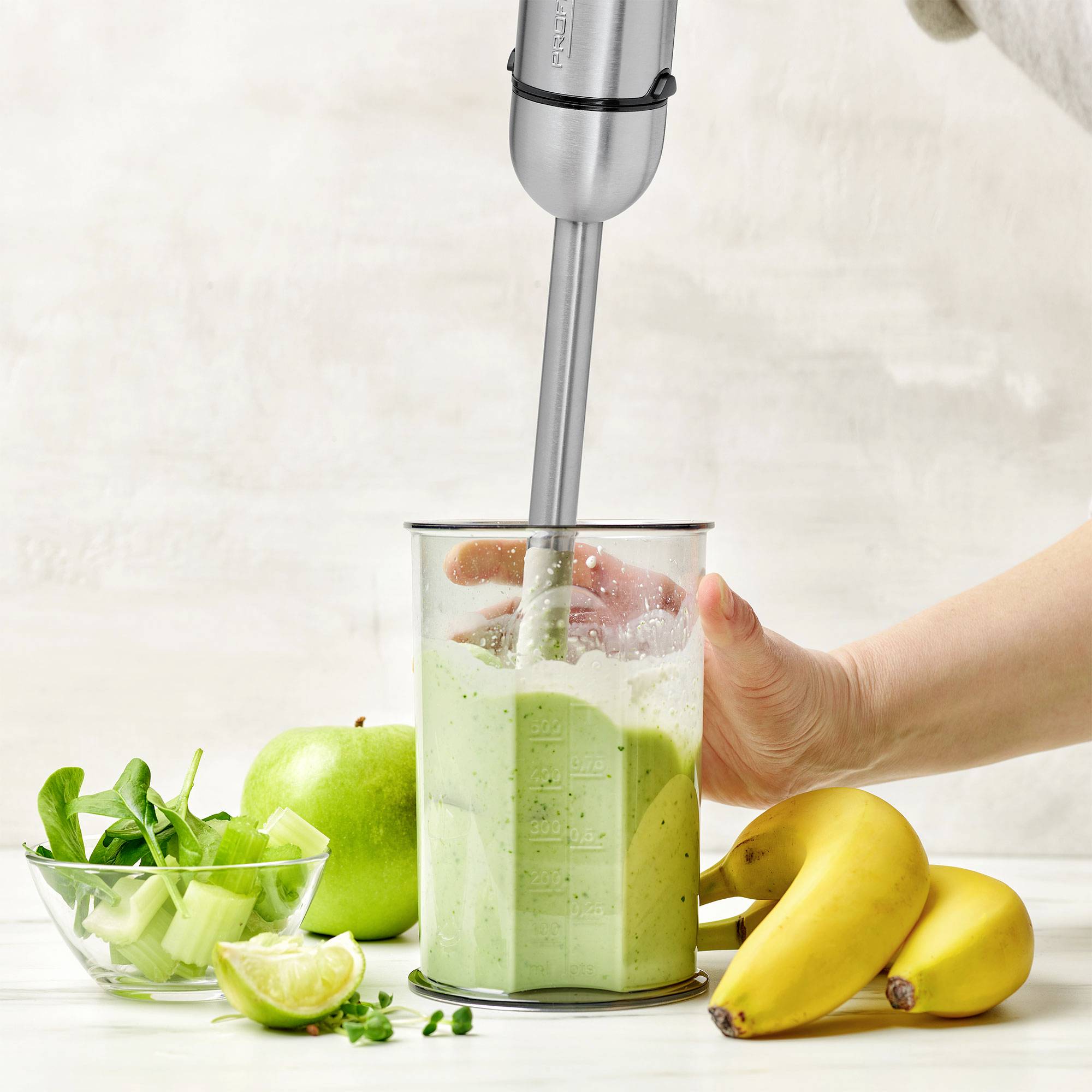 Een hand houdt een staafmixer in een glazen kom met een groene smoothie; ernaast liggen bananen, een appel en verse spinazieblaadjes.