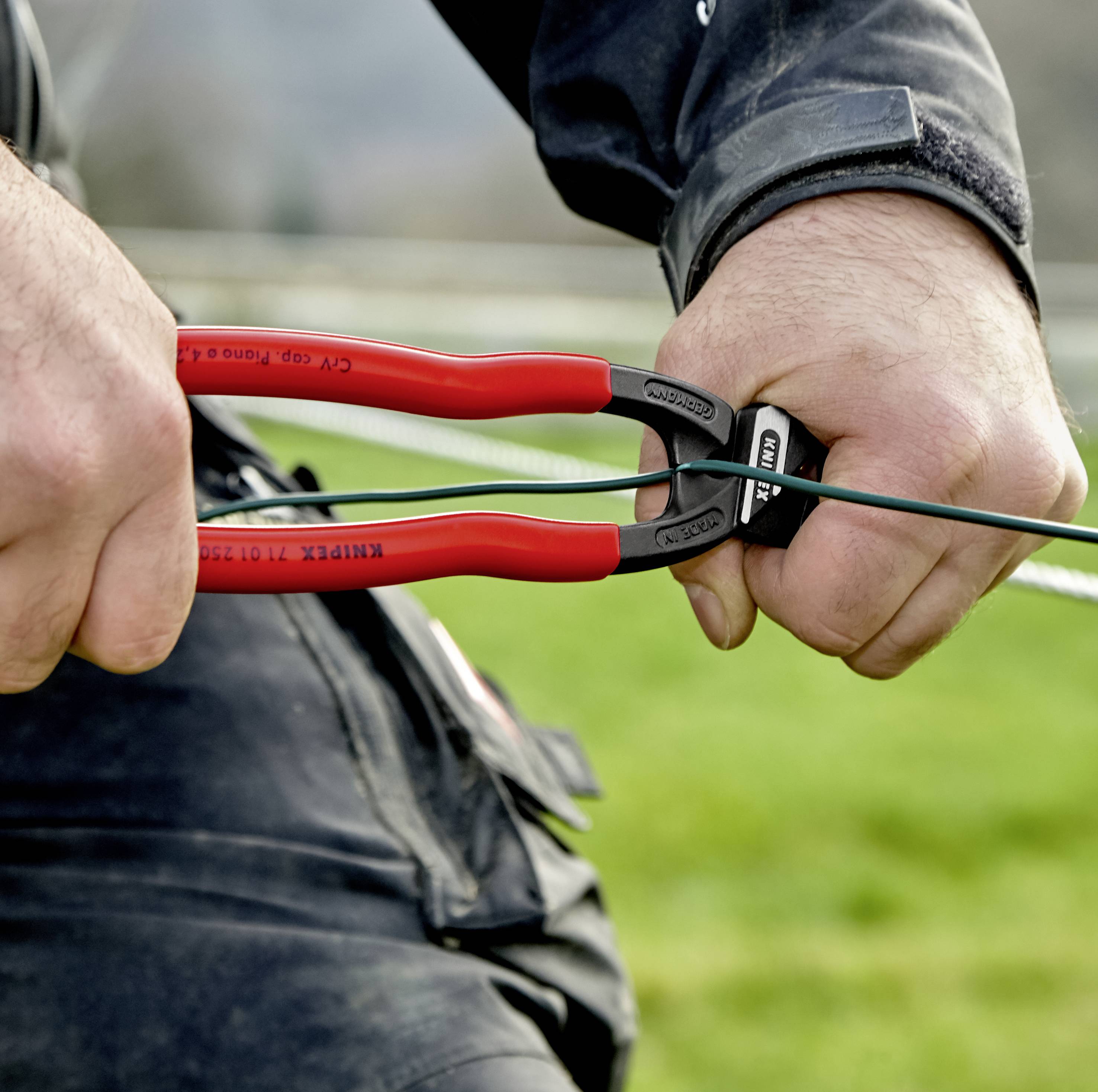 Een persoon knipt een draad door met een rode zijkniptang buiten. De handen houden het gereedschap stevig vast.