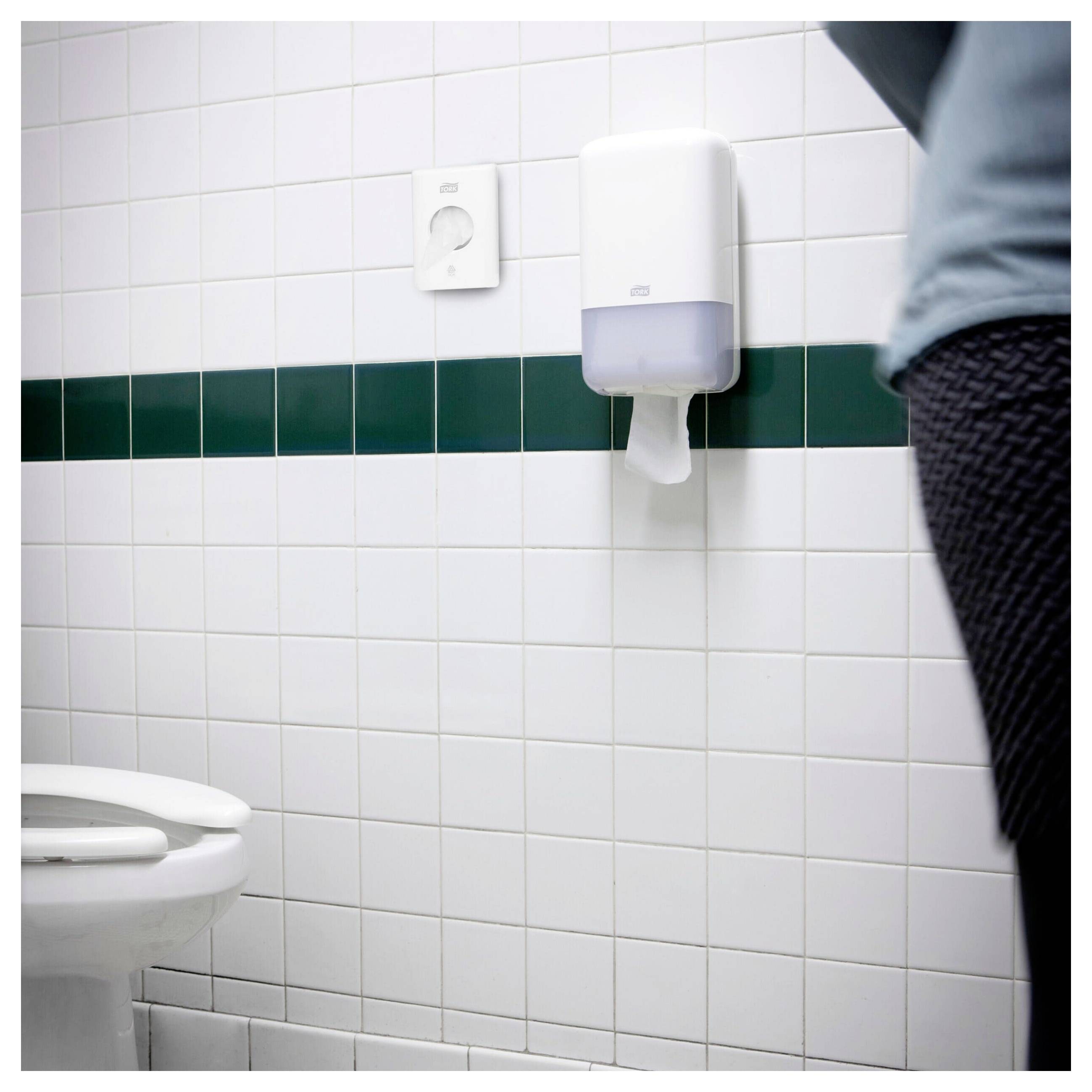 Openbaar toilet met betegelde muur, toilet en aan de muur gemonteerde zeep- of desinfectiedispenser; aan de rechterkant is een persoon gedeeltelijk zichtbaar.