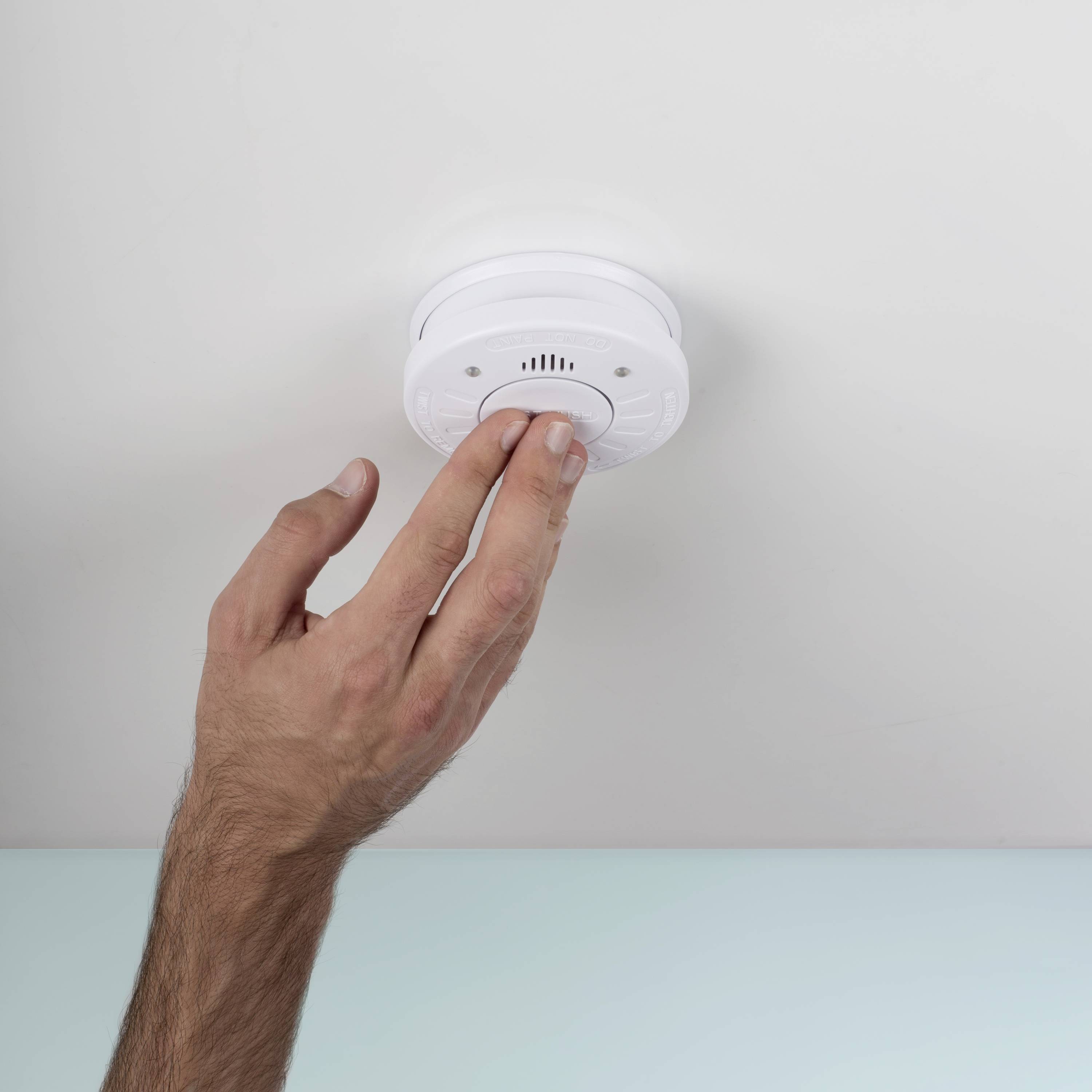 Een hand test een rookmelder aan het plafond. De melder is rond en wit, belangrijk voor veiligheid binnenshuis.