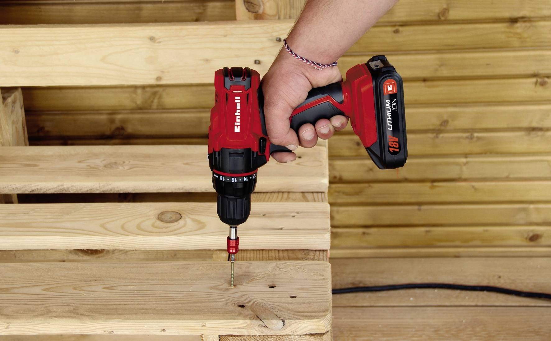 Een persoon bevestigt een schroef in een houten plank met een rode accuschroevendraaier, met op de achtergrond een houten beschot.