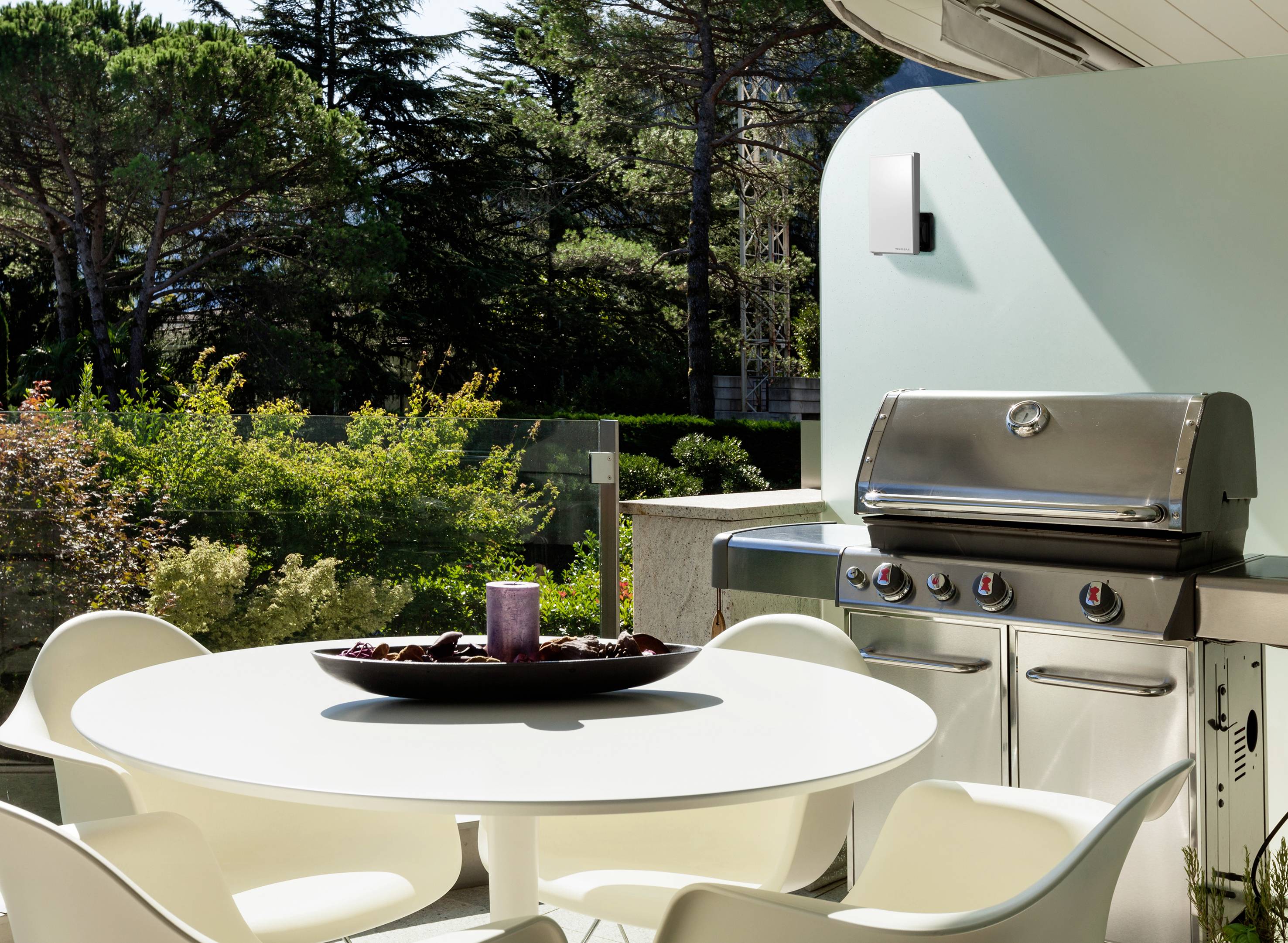 Een moderne barbecue staat op een terras met uitzicht op een tuin. Naast de barbecue staat een ronde tafel met witte stoelen.