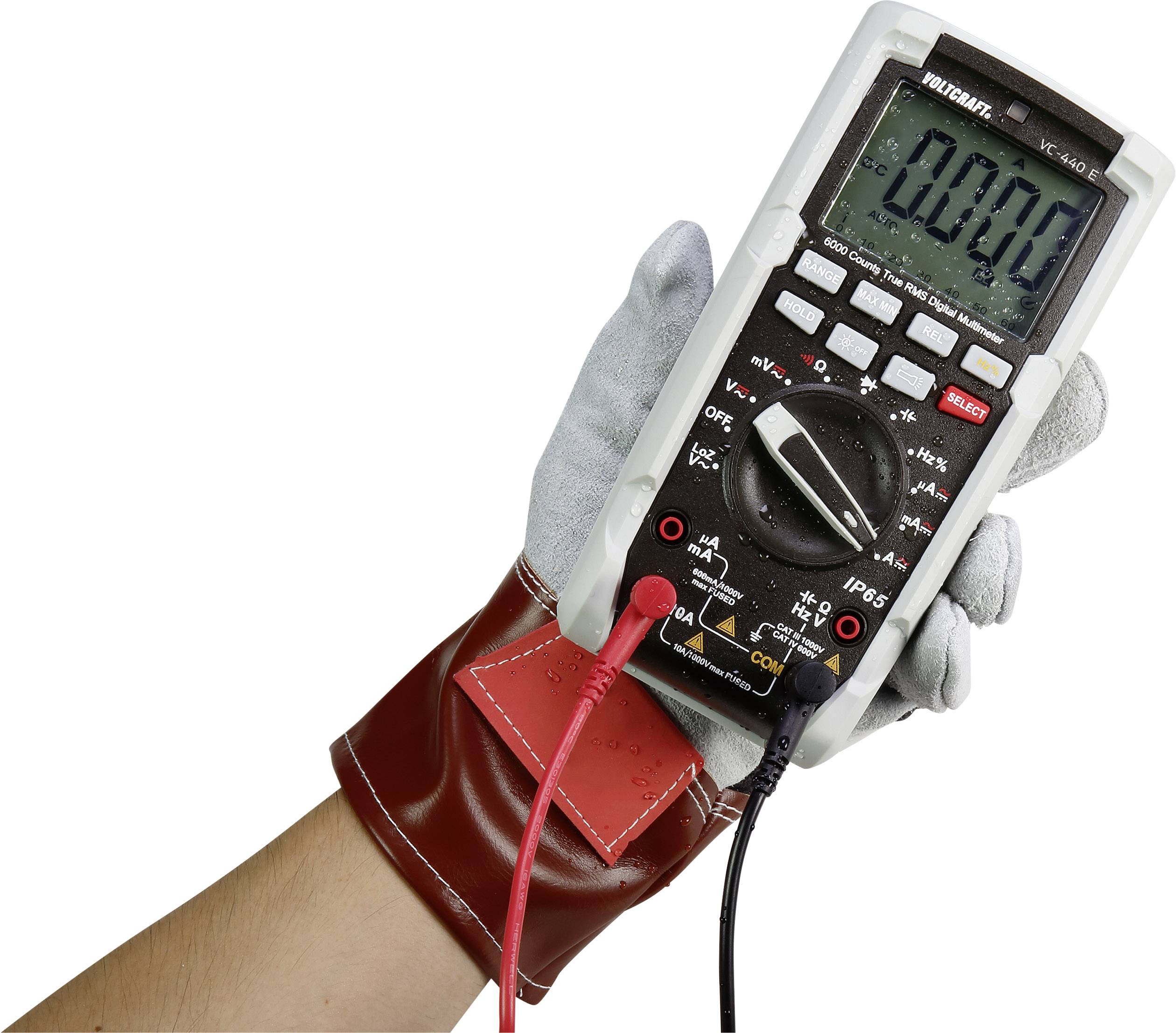 Een hand in een beschermende handschoen houdt een digitale multimeter vast met aangesloten rode en zwarte meetkabels.