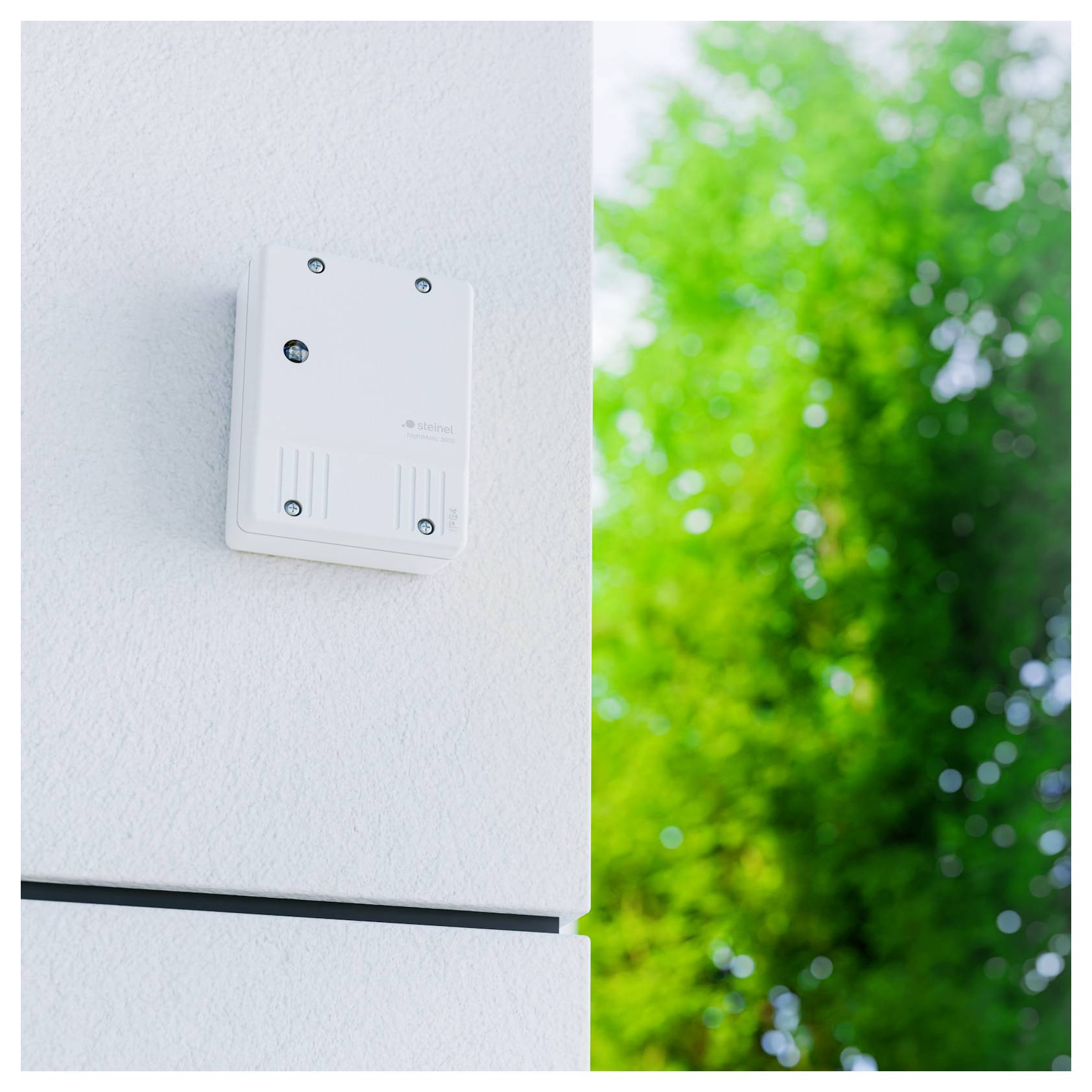 Witte aan de muur gemonteerde sensor of bedieningskast op de buitenkant van een gebouw, met een wazig groene boom op de achtergrond.