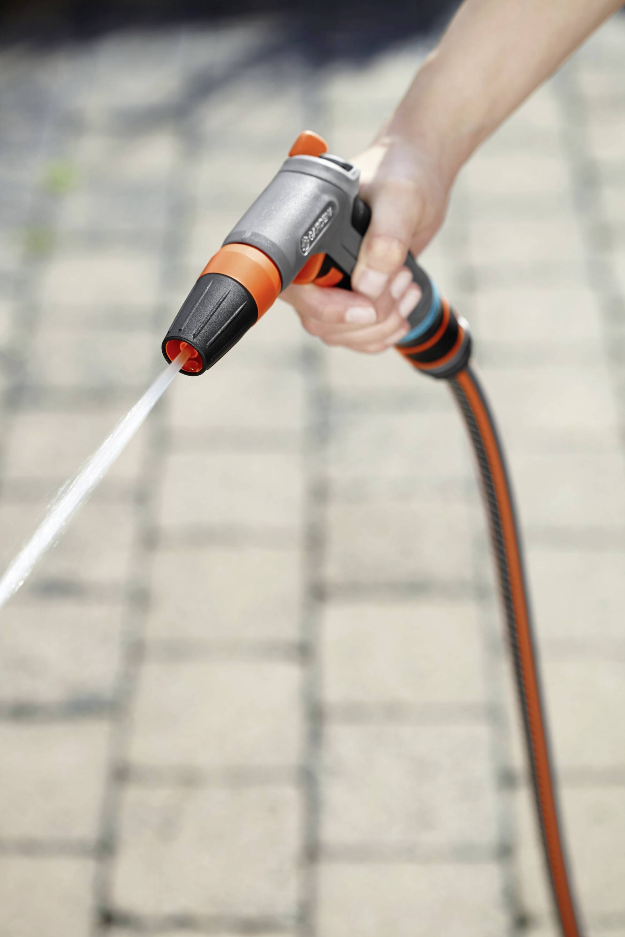 Een hand houdt een tuinslang met een sproeikop, waterstraal spuit op een bestrate ondergrond.