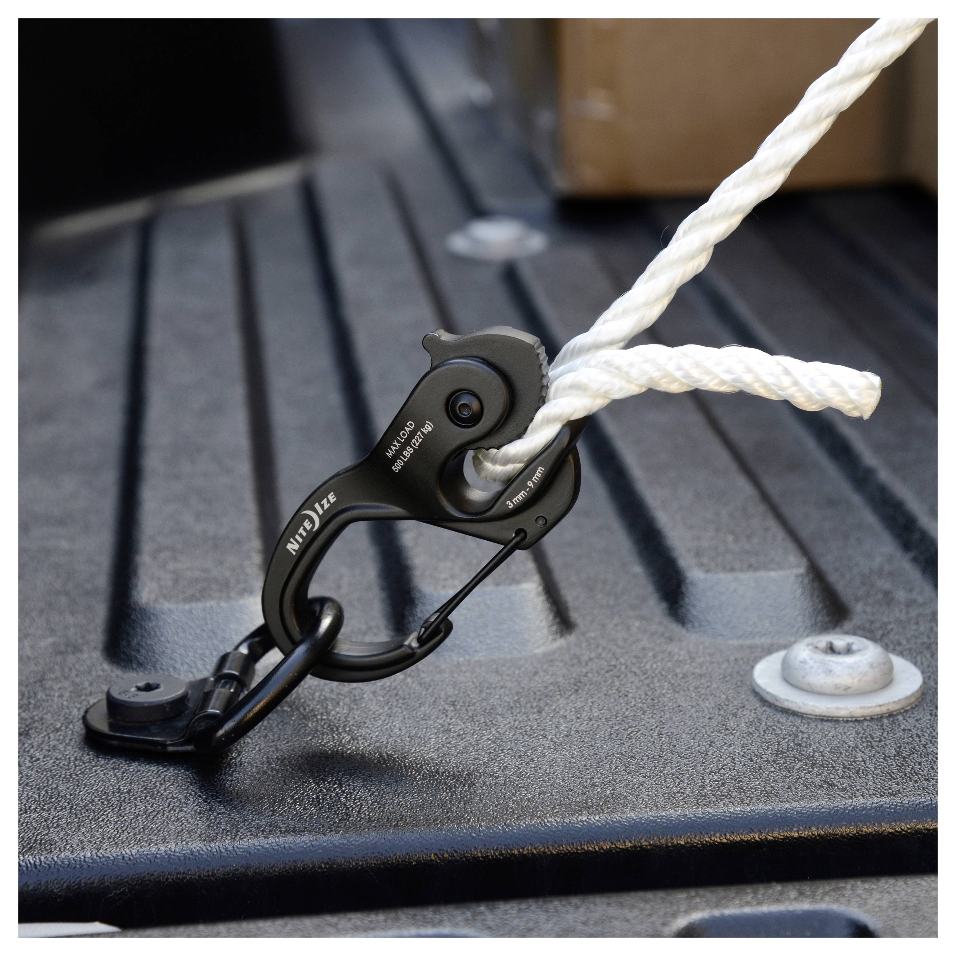 Een metalen karabijnhaak bevestigt een dikke witte touw in de laadruimte van een vrachtwagen, waarbij een methode wordt geïllustreerd voor het stevig vastmaken van lading.