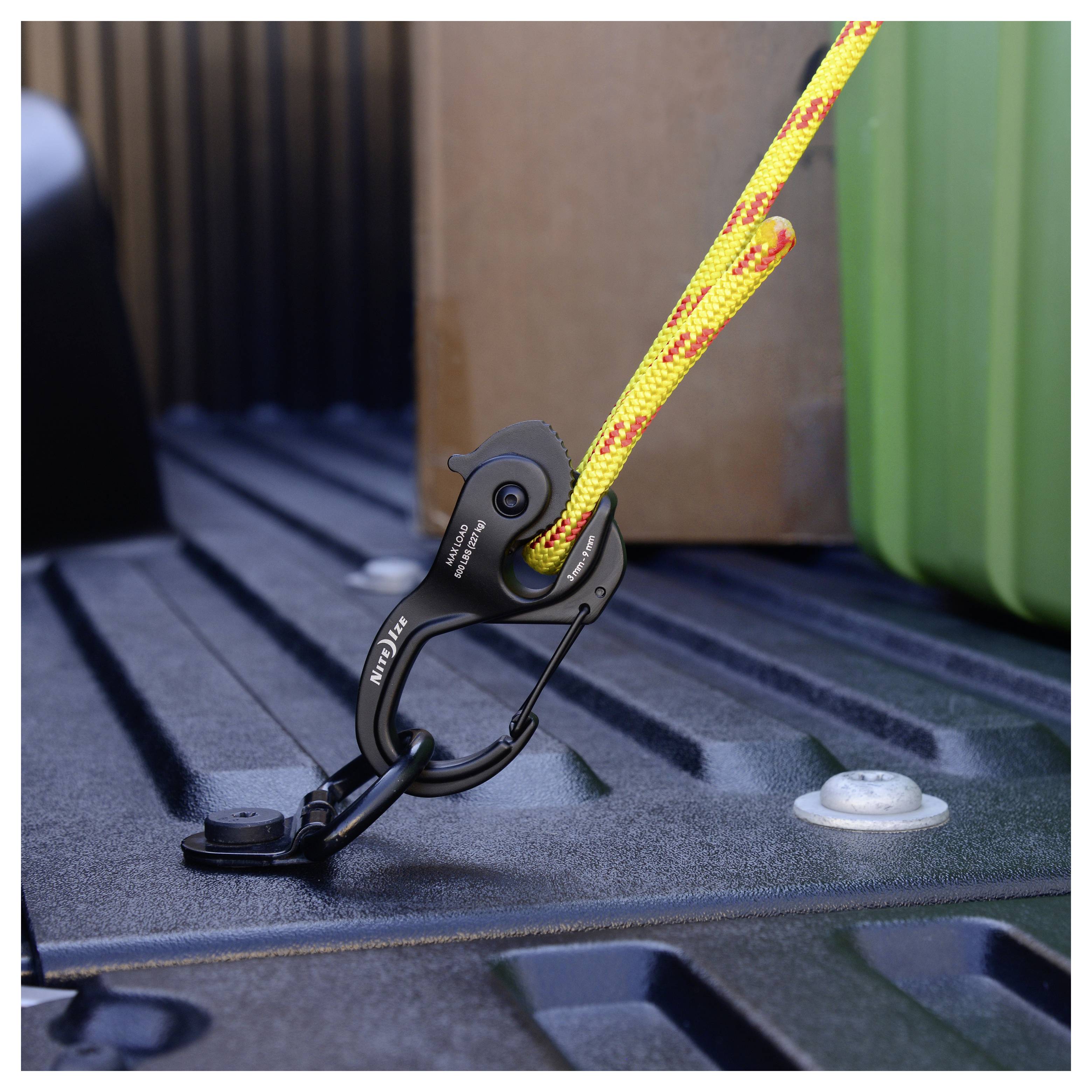 Een close-up van een zwarte klem die een geel-oranje touw bevestigt aan de laadruimte van een voertuig, naast een groen object, waarschijnlijk een container.
