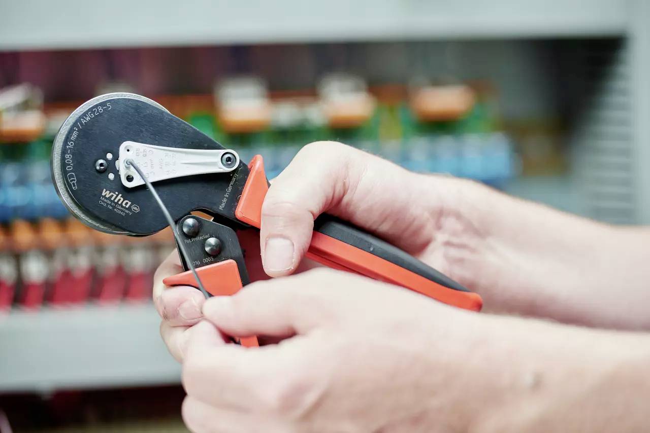 Handen houden een krimptang vast om een kabel te bewerken, met elektronische componenten onscherp op de achtergrond.