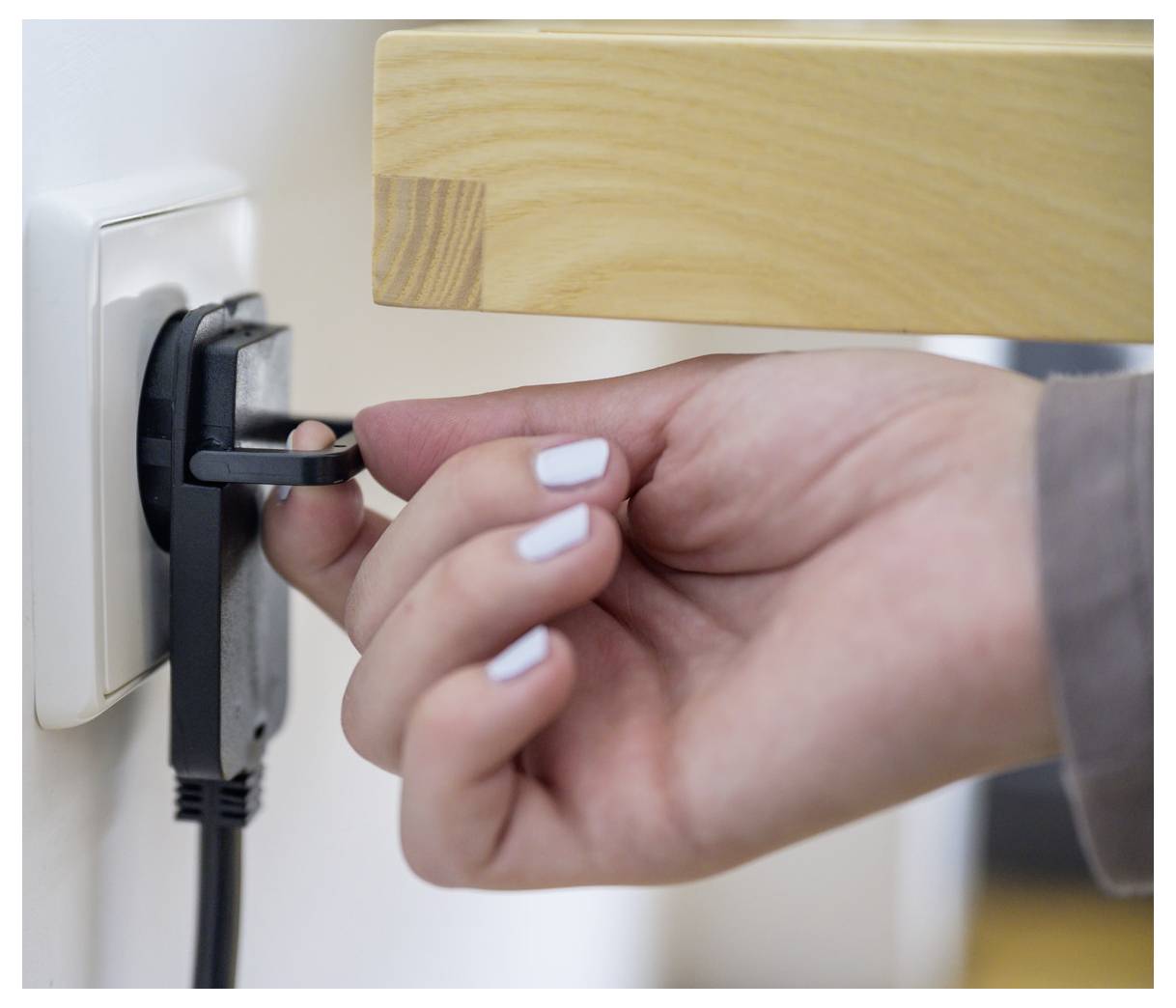 Hand die een zwarte stroomkabel in een wandcontactdoos naast een houten bedframe stopt.