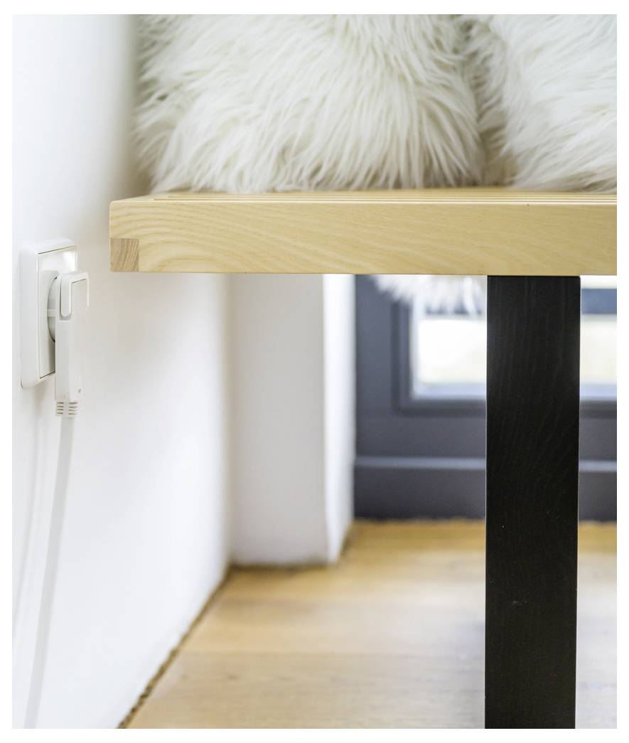Close-up van een wit zacht kussen op een licht houten bank naast een wandcontactdoos en raam, met een minimalistische binnenhoek.