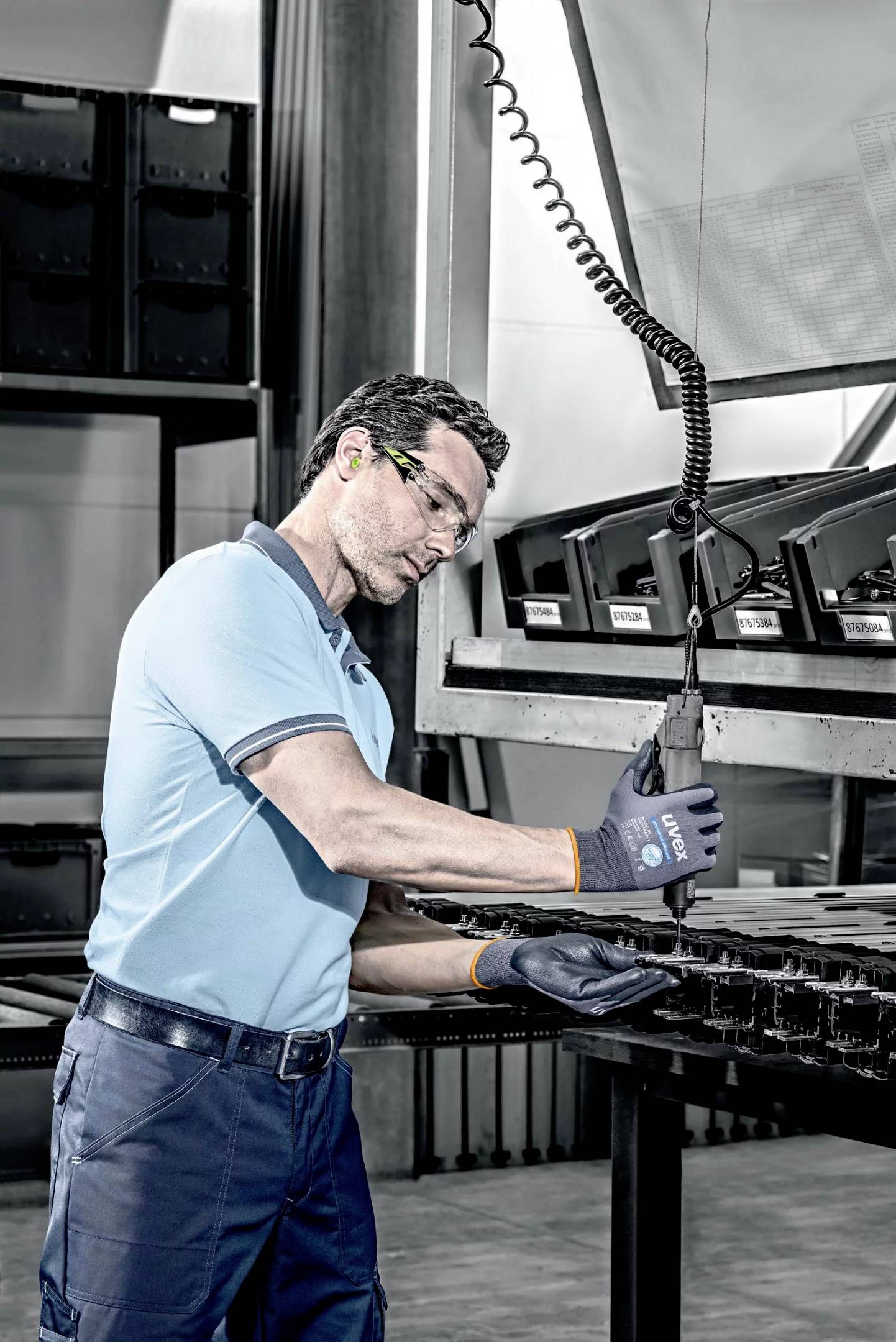 Een man in beschermende kleding werkt in een fabriekomgeving met een gereedschap aan een metalen tafel. Op de achtergrond zijn rekken te zien.