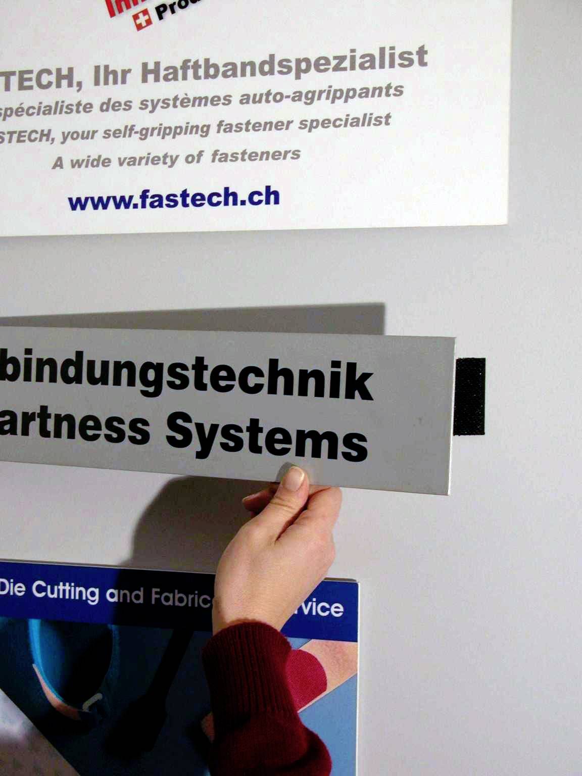 Een persoon houdt een bord vast met de tekst 'Verbindingstechniek' voor een muur waarop reclameborden van FASTECH te zien zijn.