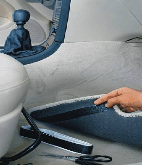 Een persoon houdt een stuk tapijt in het interieur van een auto omhoog om daaronder een kabelbinder of klittenband aan te brengen.
