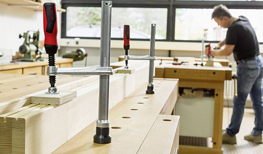 Een ambachtsman werkt in een werkplaats aan een houten tafel waarop verschillende klemmen zijn bevestigd. Op de achtergrond zijn machines zichtbaar.