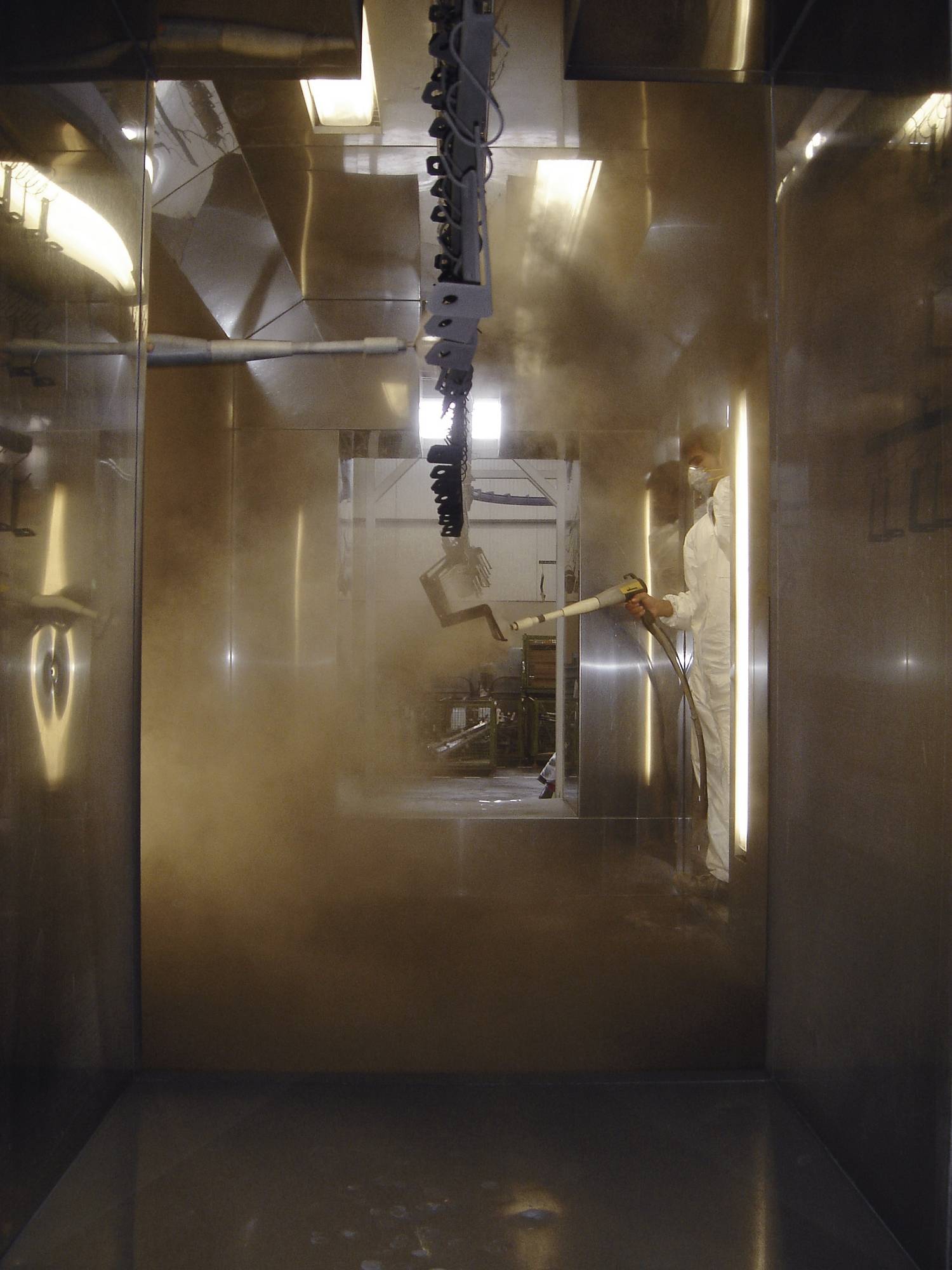 Een arbeider in beschermende kleding spuit verf in een spuitcabine. De omgeving is industrieel met metalen wanden.