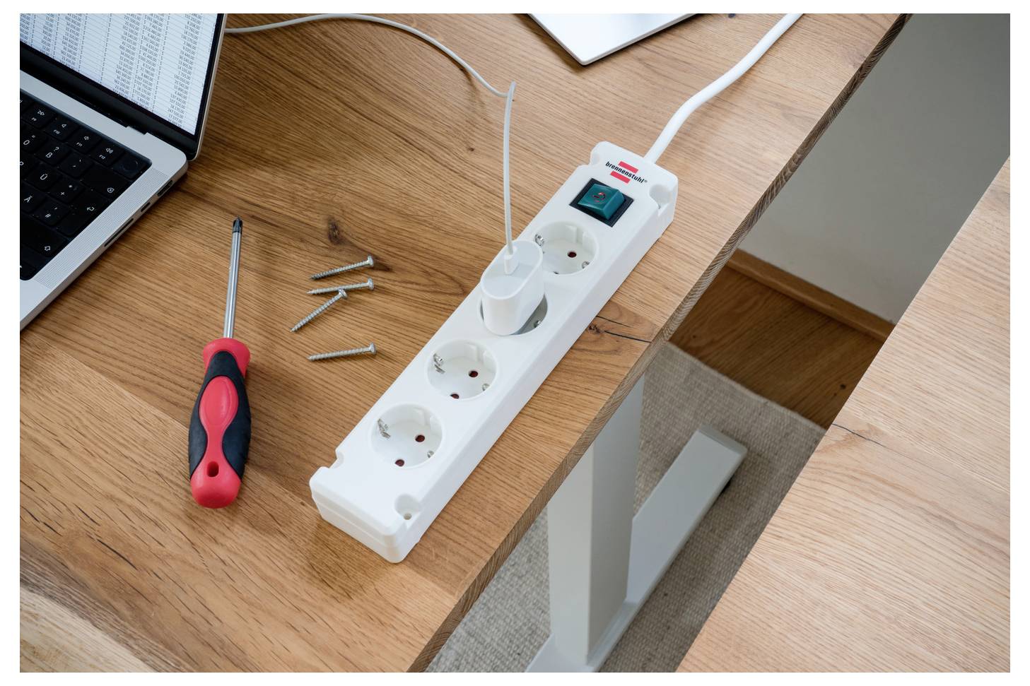 Bureau met een stekkerdoos, schroevendraaier, schroeven en een beker met gereedschap naast een laptop, wat duidt op elektrische installatie of reparatiewerkzaamheden.