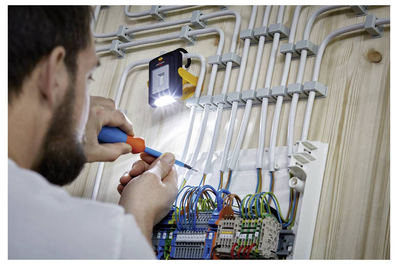 Elektricien die een elektrisch paneel bedraden, gekleurde draden in een aan de muur gemonteerde distributiebak aansluitend terwijl hij een zaklamp voor zichtbaarheid gebruikt.
