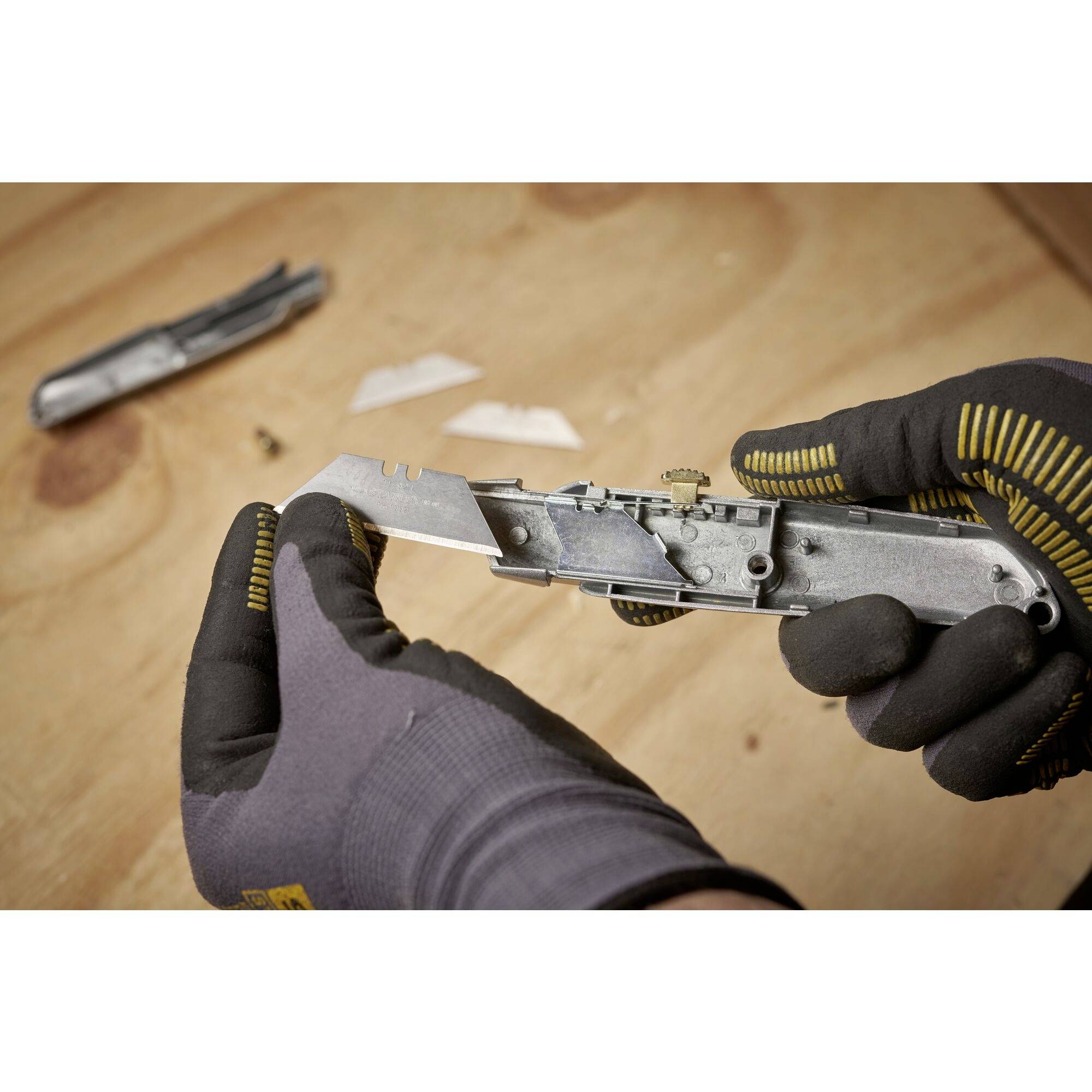 Een persoon met een grijze handschoen houdt een zilveren gereedschap vast op een houten tafeloppervlak. Op de achtergrond liggen nog meer metalen onderdelen.