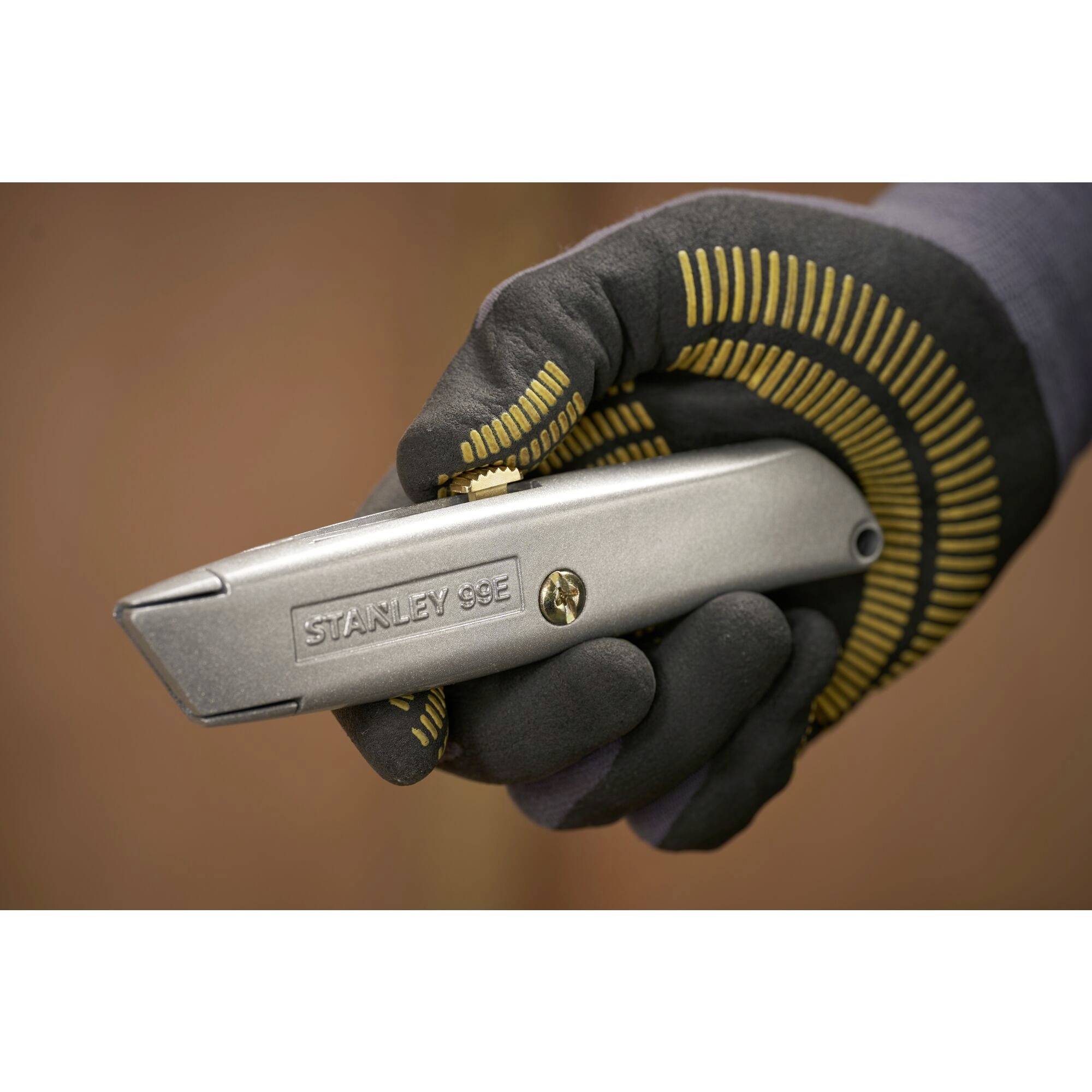 Een aangeklede hand houdt een metalen cuttermes vast. Het mes draagt de opdruk 'Stanley 99E' en beschikt over een schroefmechanisme.