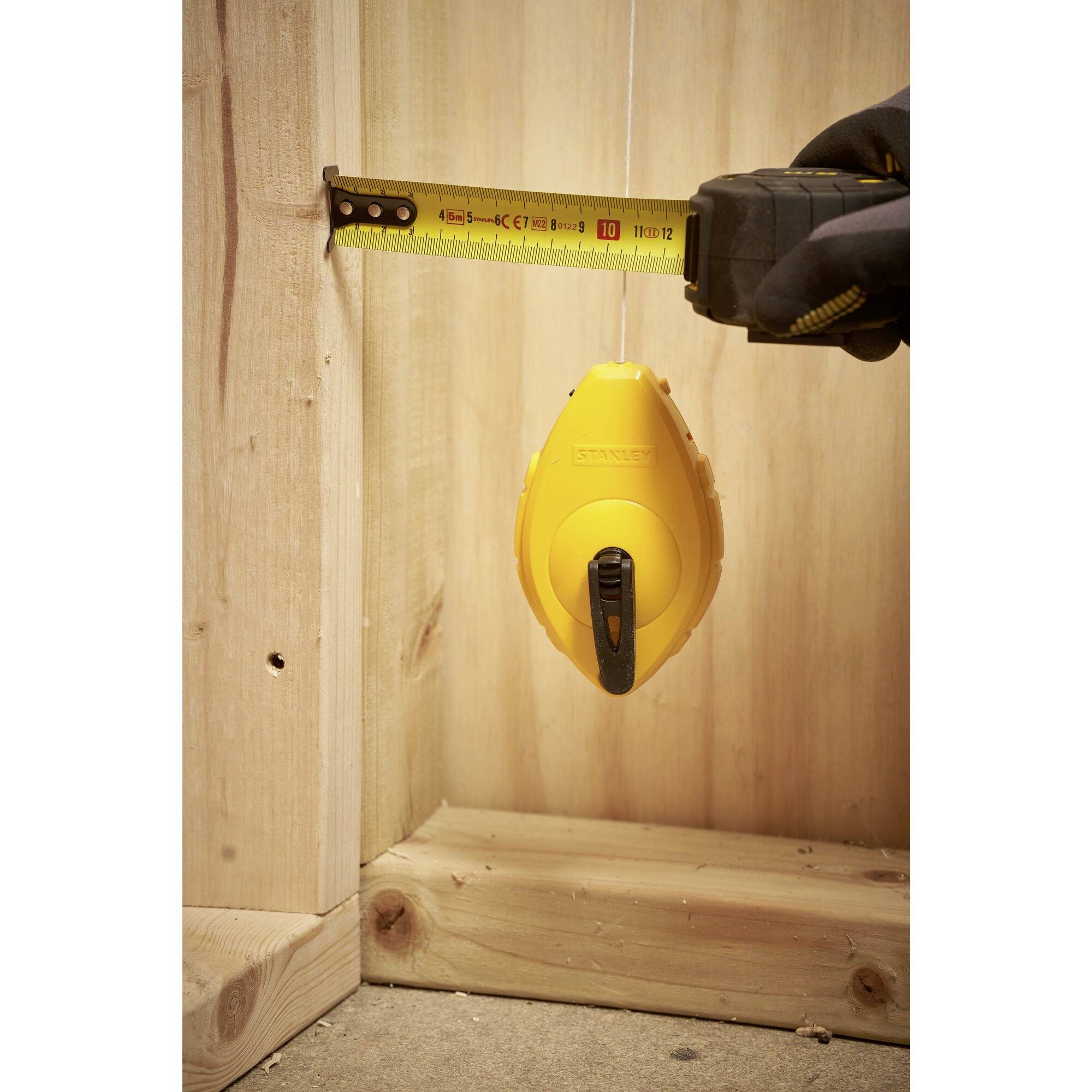 Een geel krijtlood hangt aan een wandhouder in een houten lijst, terwijl iemand links de breedte opmeet met een meetlint.