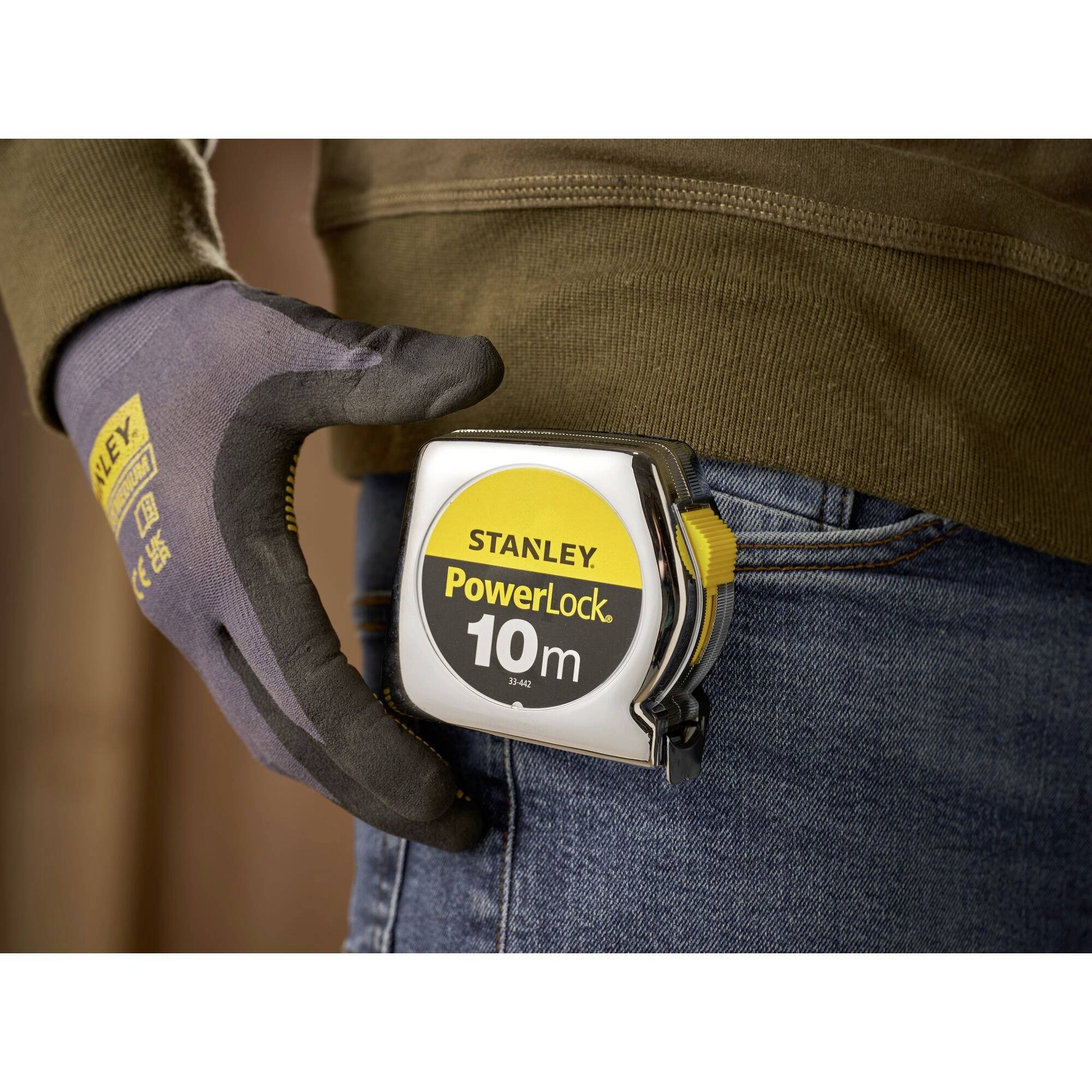 Een hand met handschoen houdt een meetlint 'Stanley PowerLock 10m' vast, dat aan een spijkerbroekrand is bevestigd.