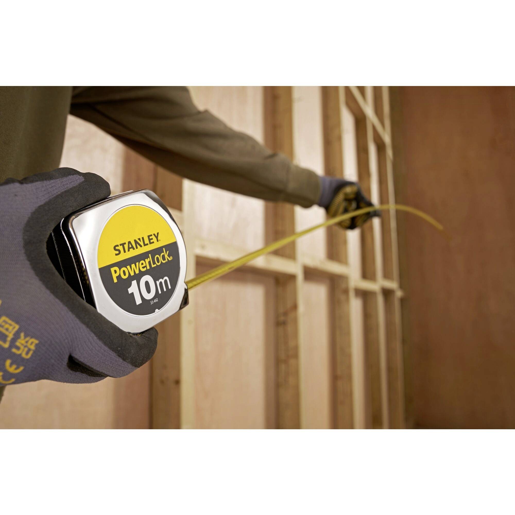 Een persoon meet een houten frame met een geel meetlint. Op het meetlint staat 'Stanley PowerLock 10m'.