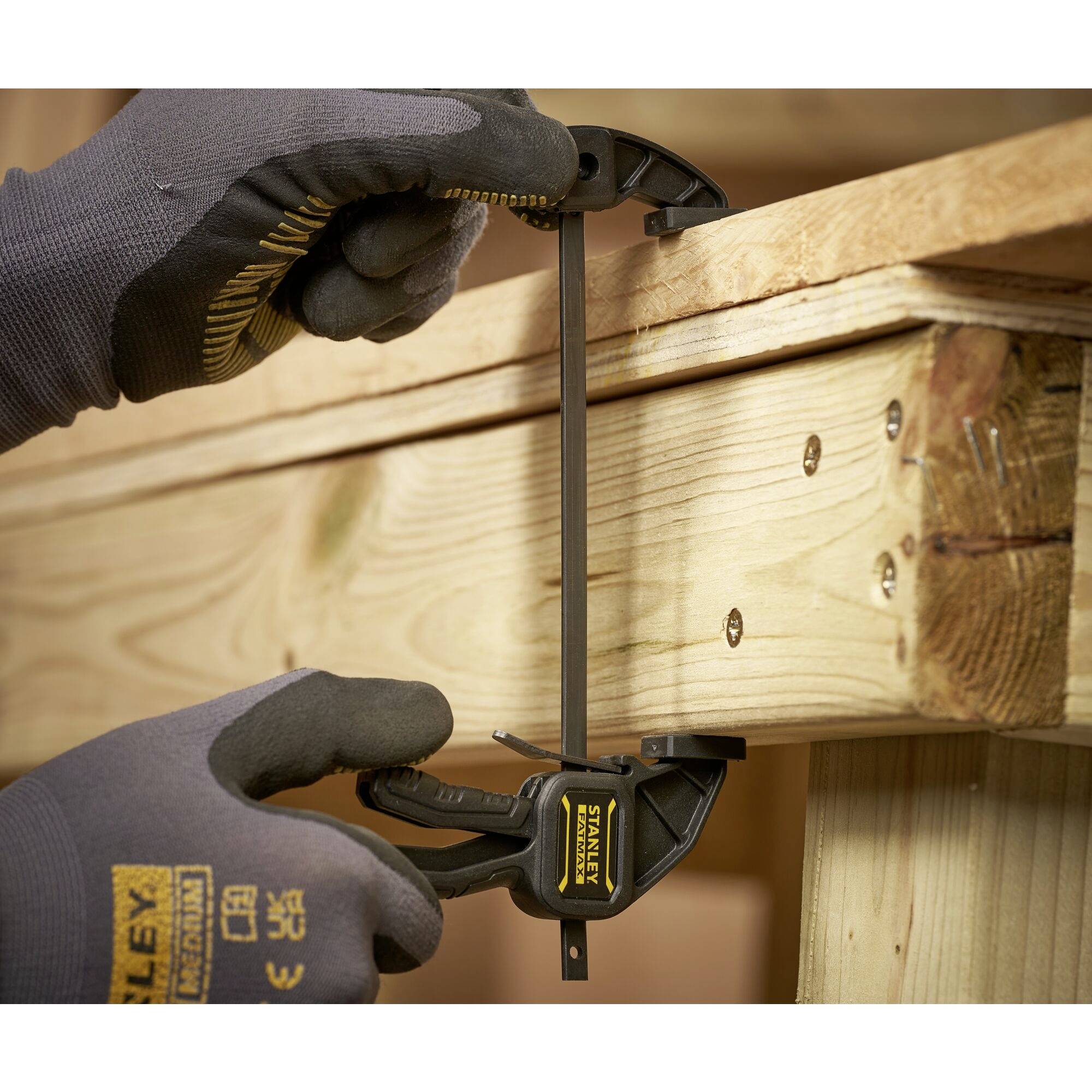Twee handen met werkhandschoenen bevestigen een houten lat met een klem aan een houten balk.