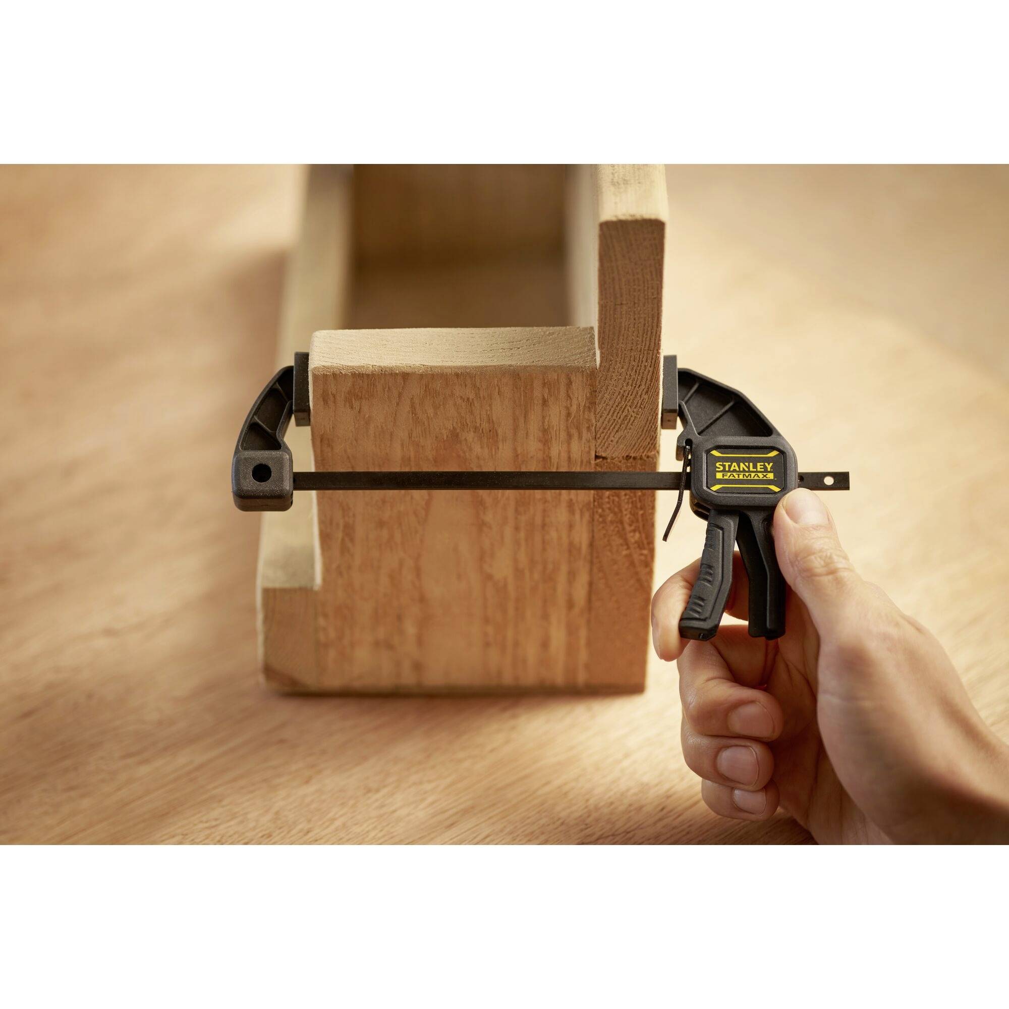 'Hand houdt klem vast, bevestigt houten stuk aan houten constructie. Gladde houten vloer. Focus ligt op het hanteren van het gereedschap.'