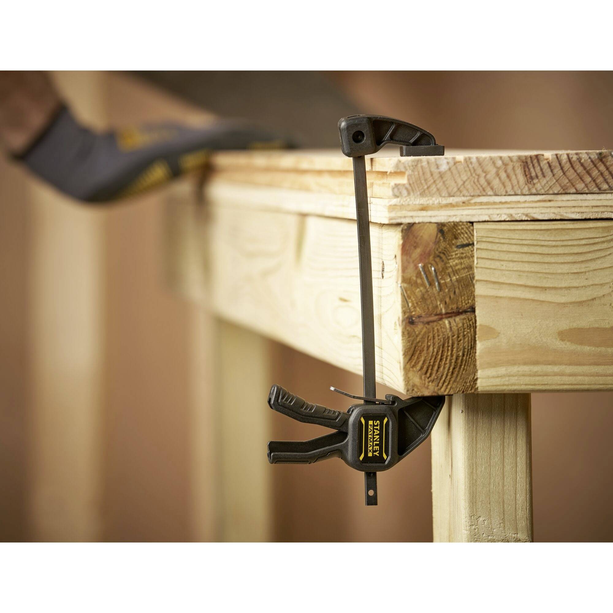 'Een klem houdt een houten stuk vast aan een werktafel. Een hand met handschoen fixeert het houten stuk. Sfeer van een werkplaats.'