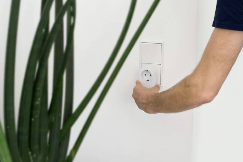 Een arm van een persoon steekt een stekker in of trekt deze uit een stopcontact aan een witte muur. Links zijn plantendelen zichtbaar.