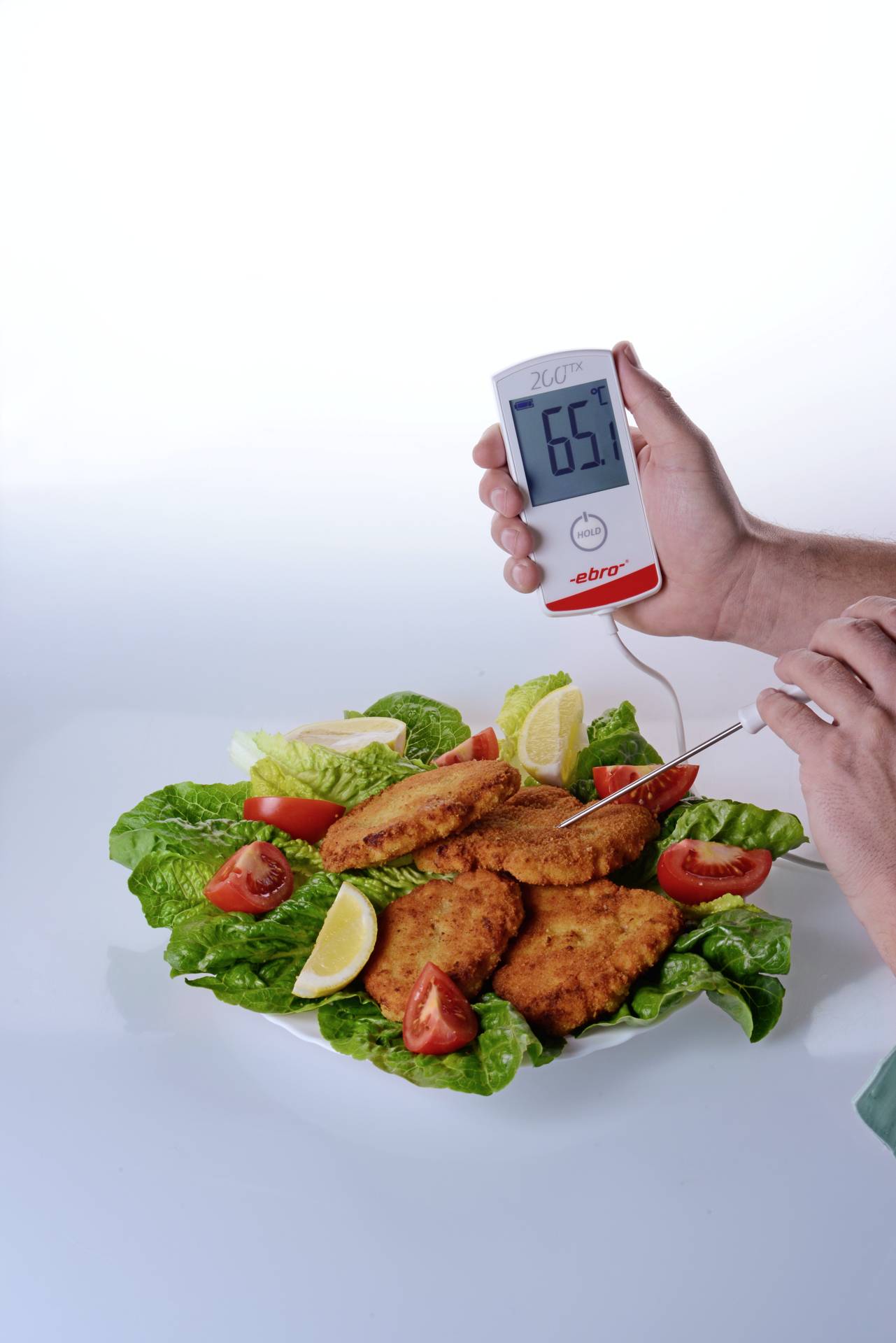 Een hand houdt een thermometer vast die in een gepaneerd kippenschnitzel wordt gestoken, liggend op een bedje van sla op een bord. Het display toont 65°C.