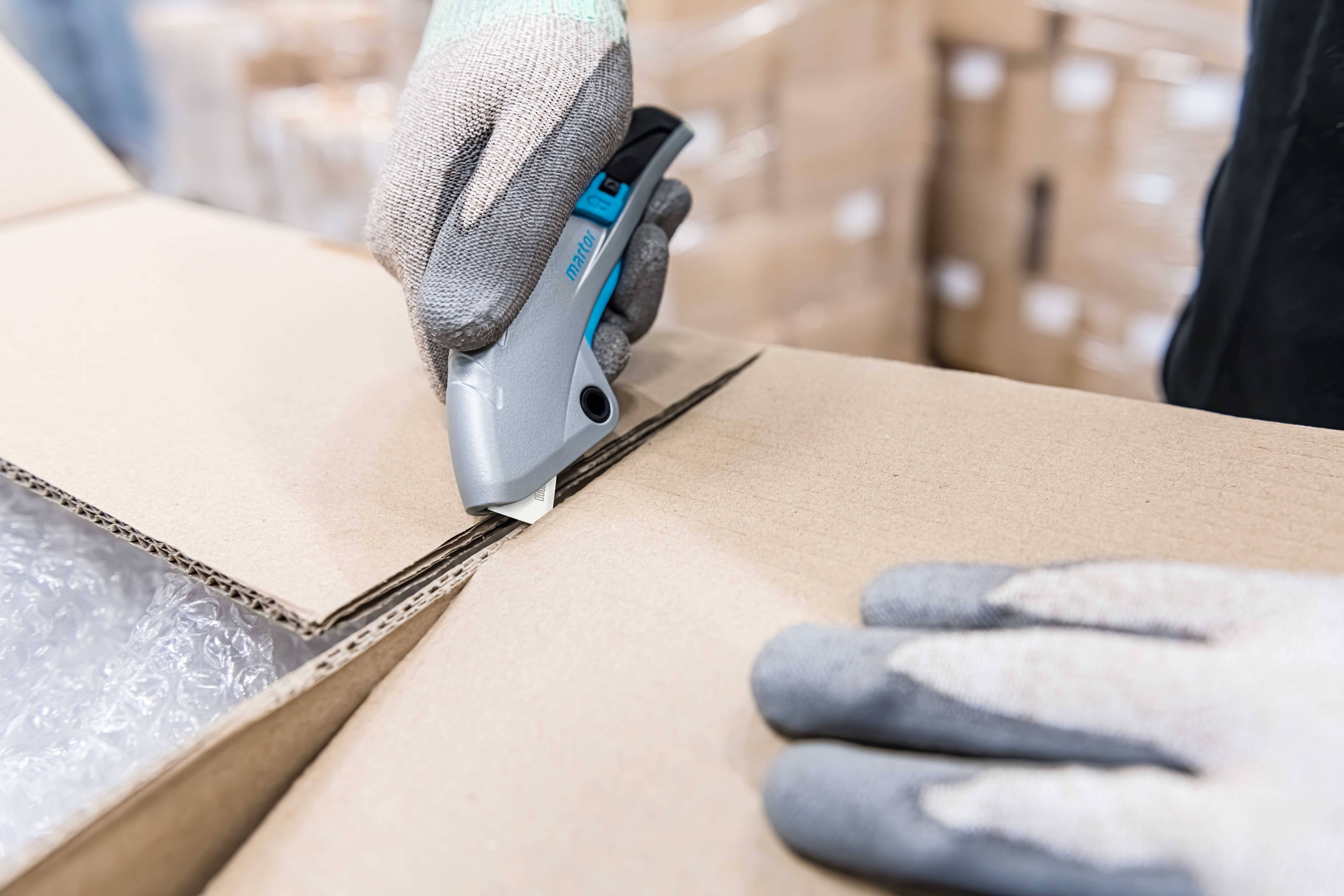 Een persoon snijdt met een grijs cuttermes een kartonnen doos. De doos bevat plastic folieverpakkingen. Handen dragen werkhandschoenen.