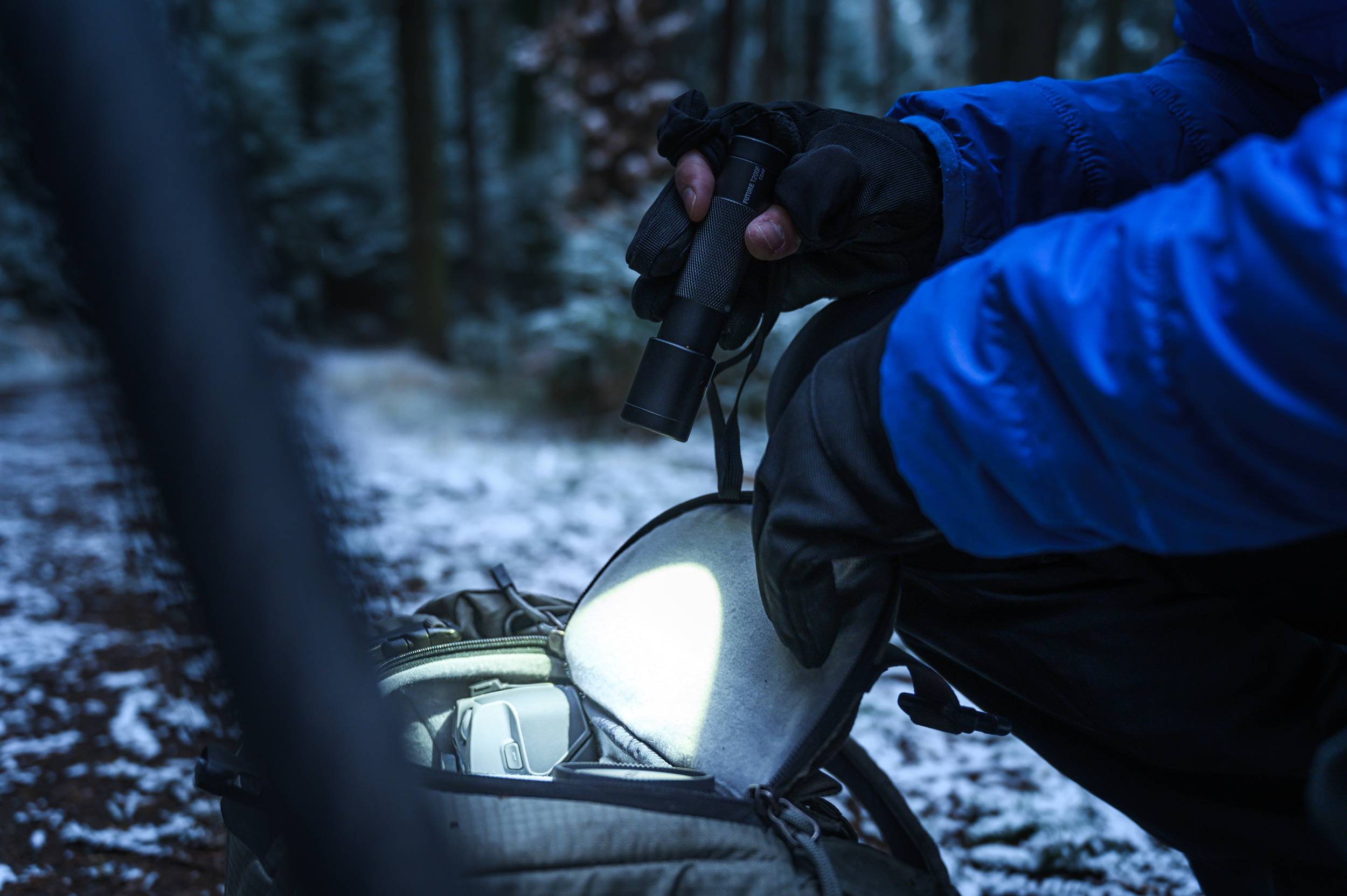 Een persoon in een winterjas houdt een zaklamp vast en doorzoekt een rugzak in een besneeuwd bos. Sneeuw bedekt de grond.