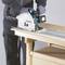 Een persoon zaagt met een elektrische handcirkelzaag een houten plank op een werktafel. De zaag is blauw en wit.