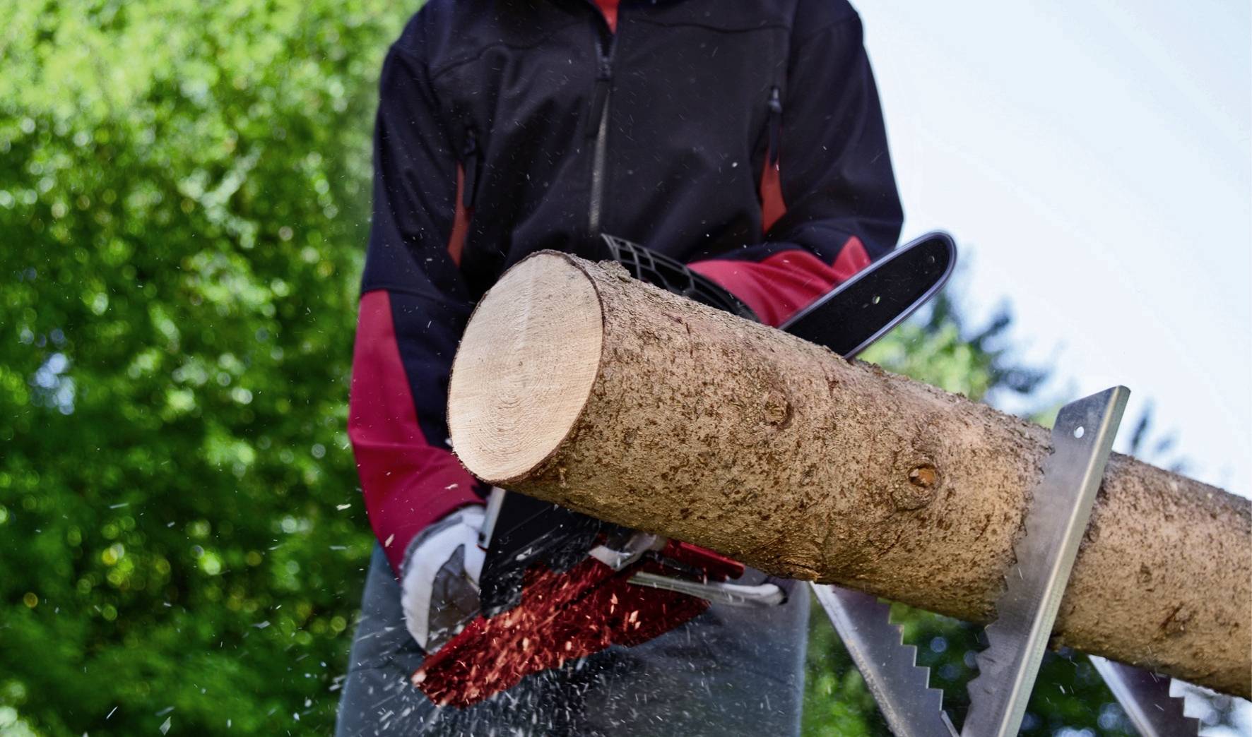 Een persoon zaagt een boomstam buiten met een motorzaag. Vonken spatten rond. Achtergrond wazig met bomen.