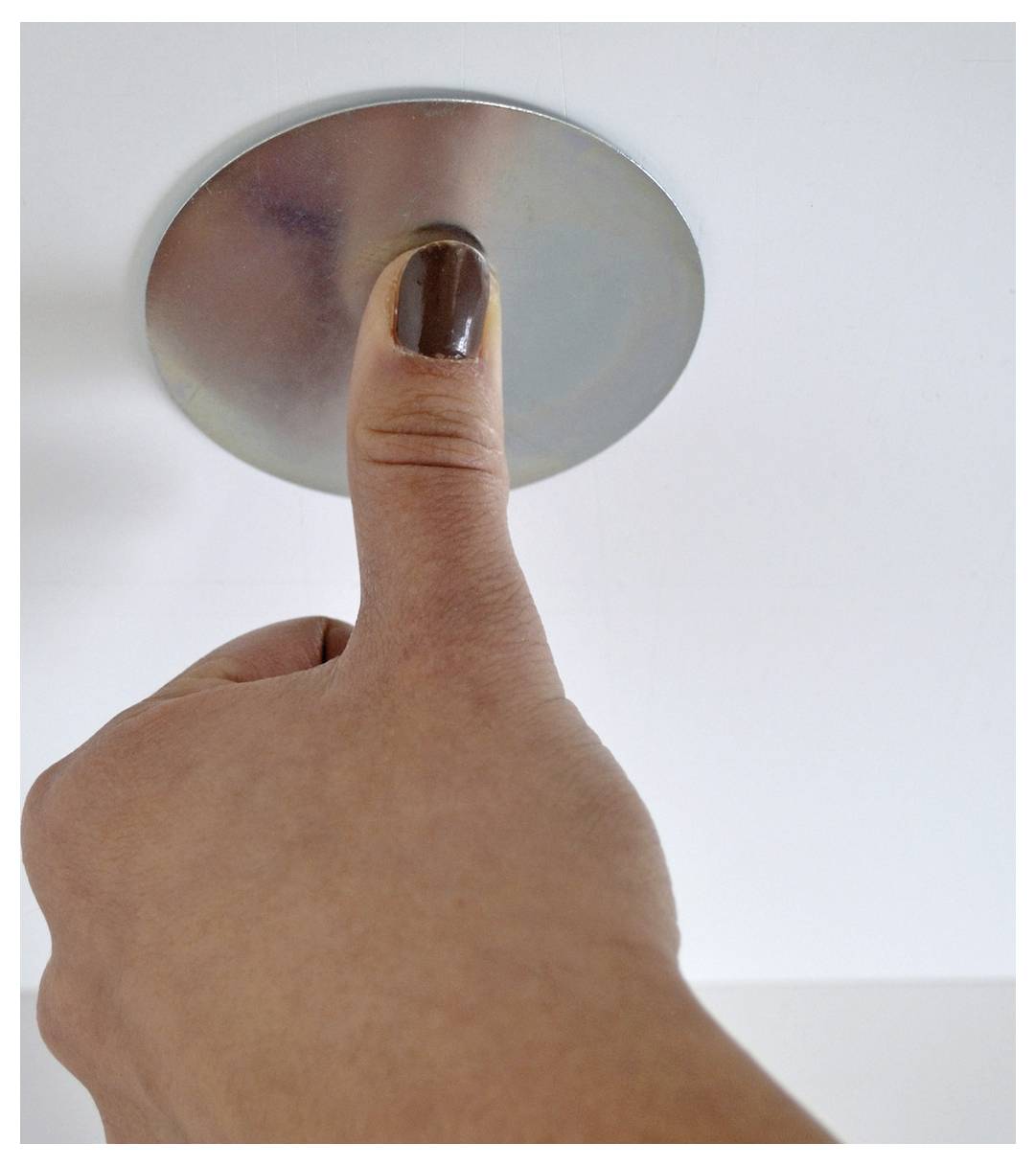 Een hand met een geschilderde duimnagel drukt op een ronde metalen knop of plafondbevestiging tegen een witte achtergrond.