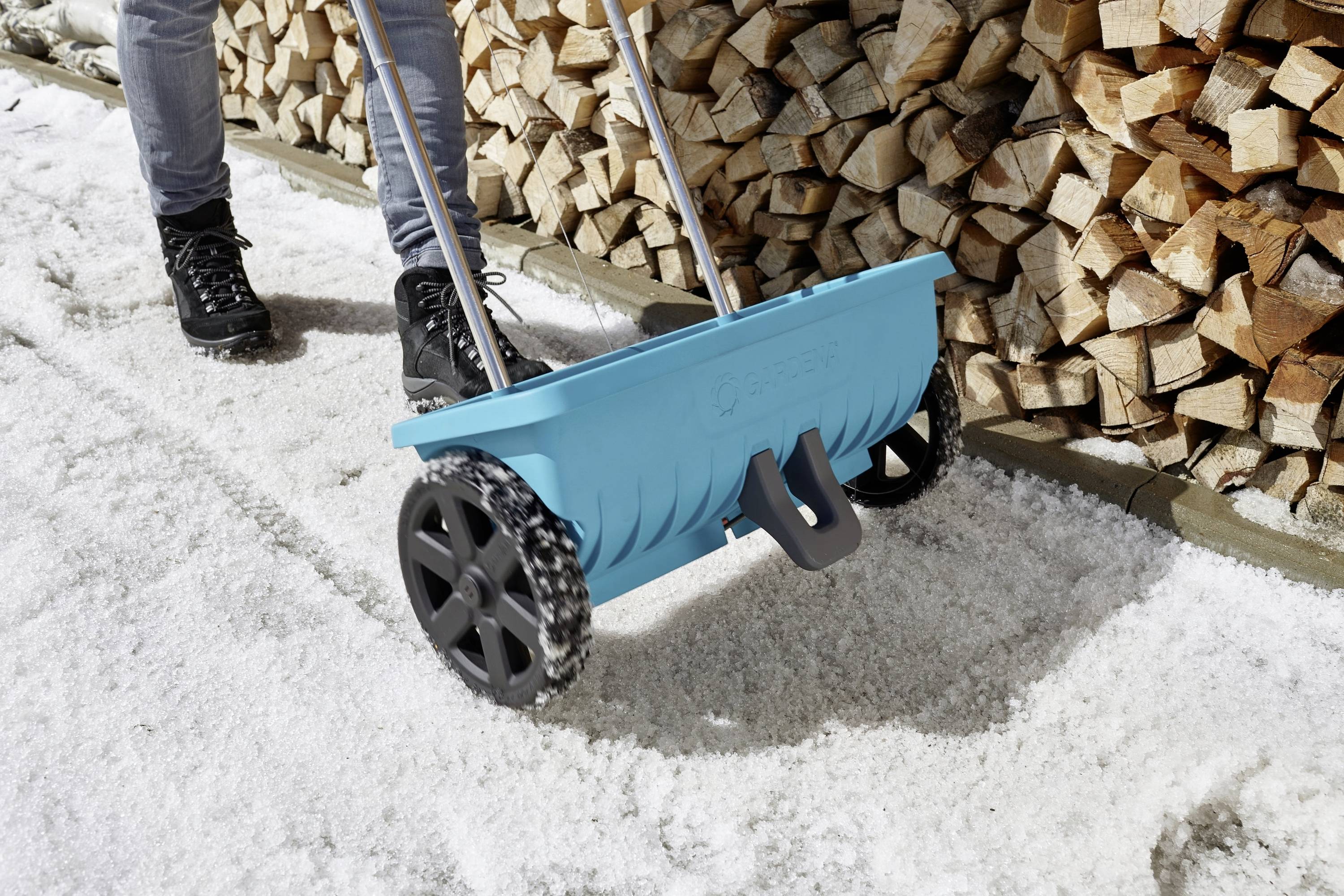 Een persoon gebruikt een blauwe strooiwagen om zout te verspreiden op een met sneeuw bedekte stoep, naast gestapeld brandhout.