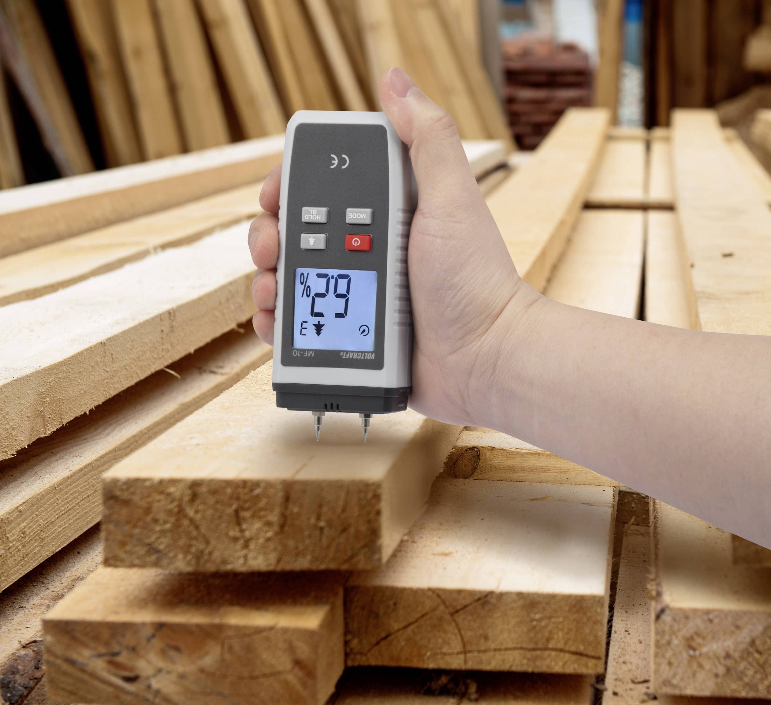 Een hand houdt een vochtmeter vast die 29% aanwijst, boven een stapel houten latten in een werkplaats.