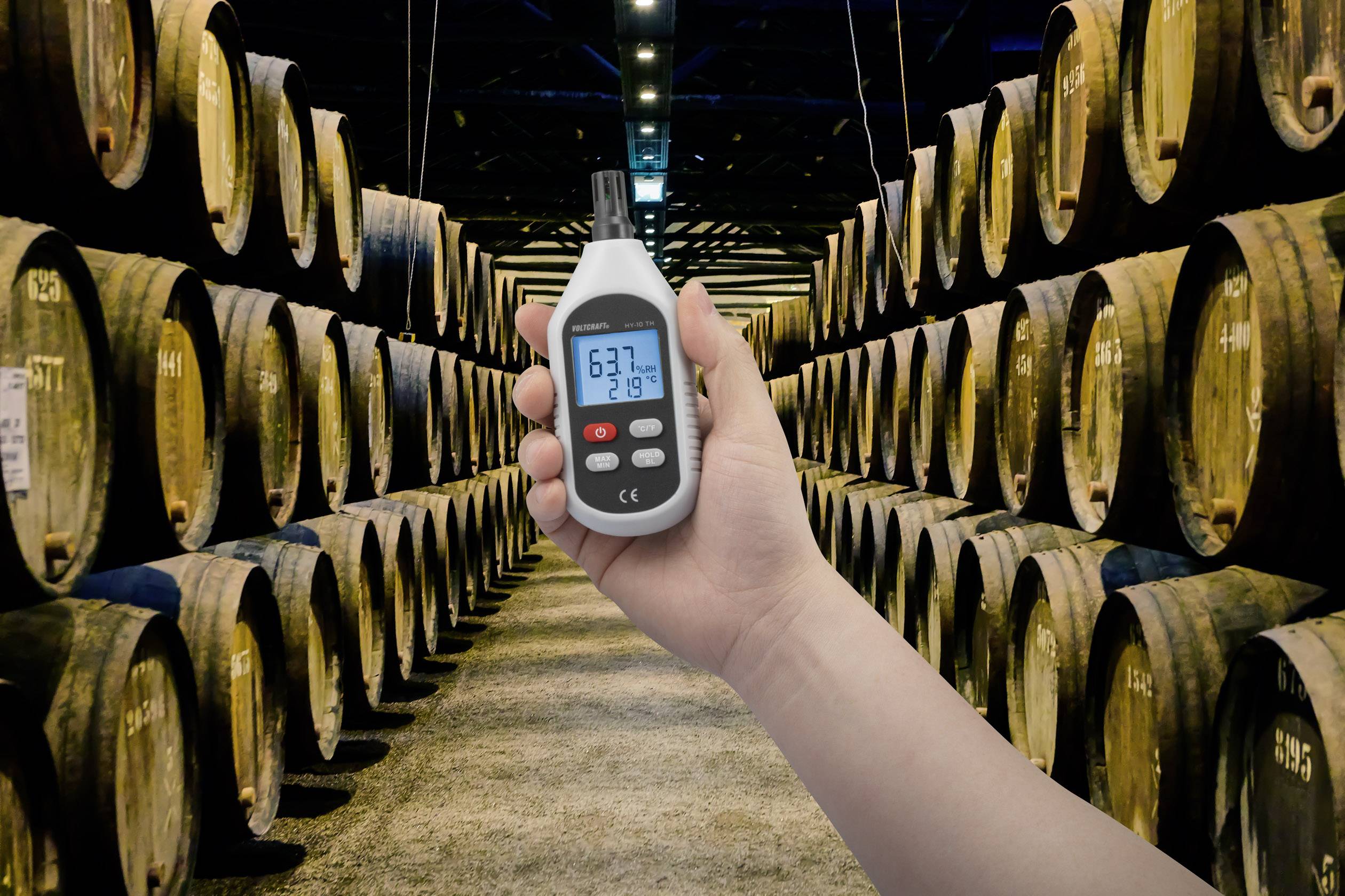 Een hand houdt een hygrometer vast die 63,7% luchtvochtigheid en 21,8 °C aangeeft. Op de achtergrond zijn houten vaten te zien in eenopslagruimte.