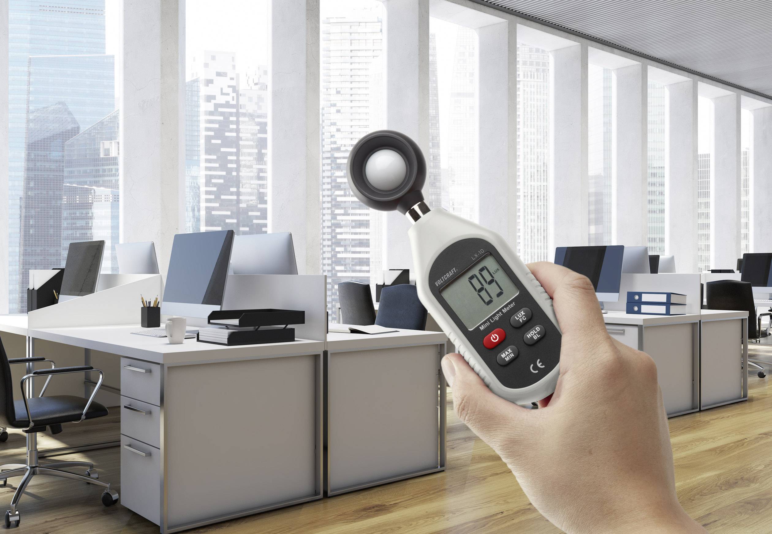 Een hand houdt een digitale thermometer vast in een modern kantoor met bureaus, computers en hoge ramen op de achtergrond.