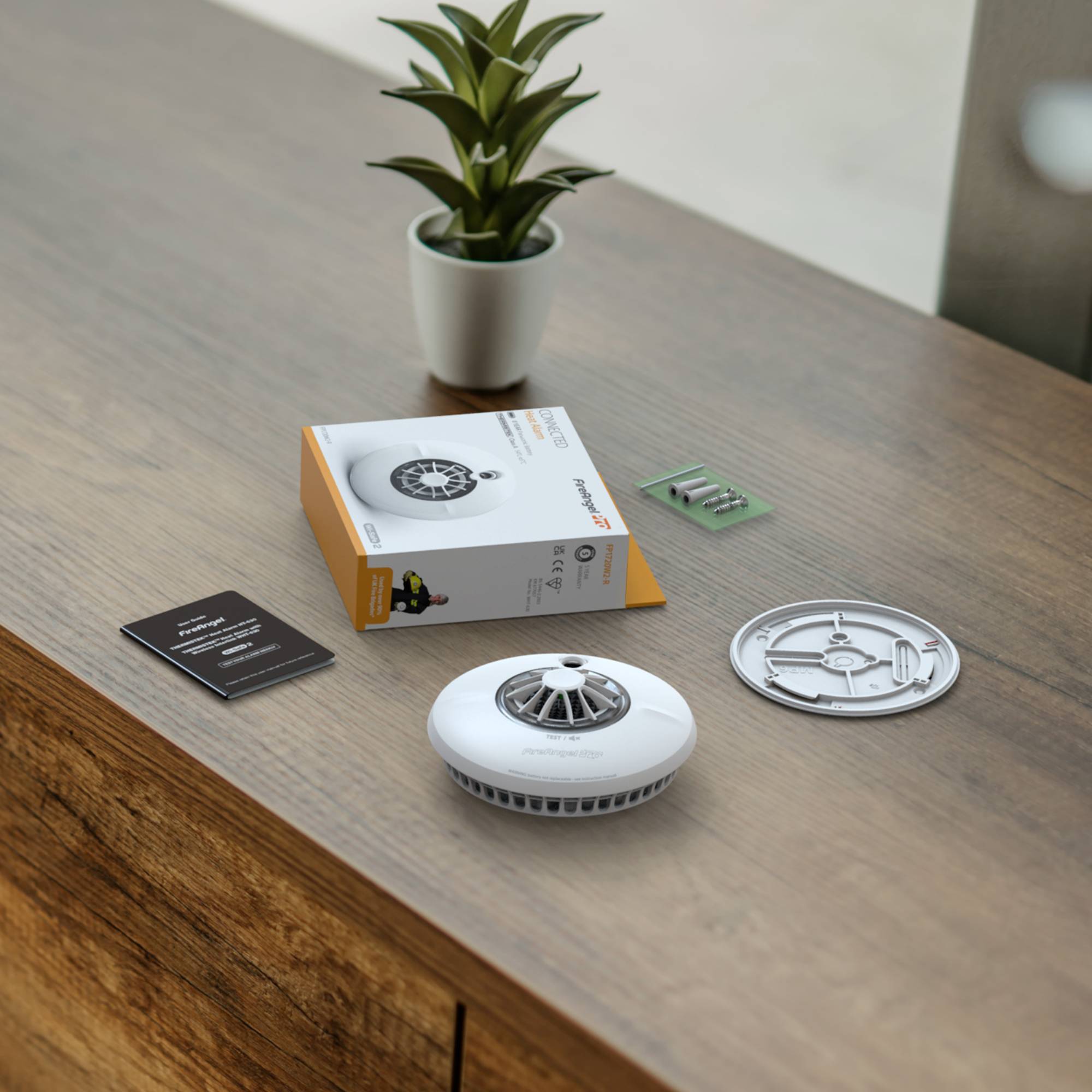 Een ronde rookmelder ligt op een houten tafel naast zijn verpakking. Op de achtergrond staat een kleine potplant.