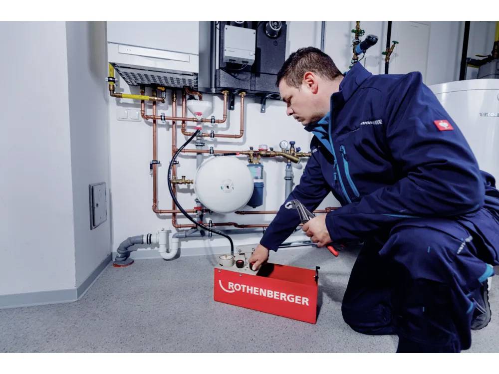 Een technicus met een blauwe overall hurkt naast een verwarmingssysteem, waarbij hij een gereedschap uit een 'Rothenberger' doos gebruikt, terwijl hij leidingen en meters inspecteert.