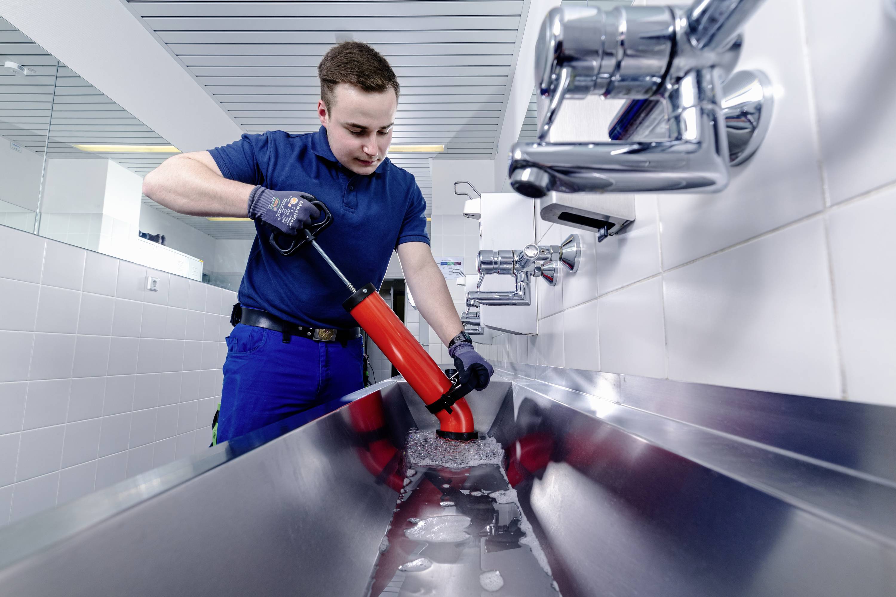 Een loodgieter in een blauwe overall werkt aan een wastafel en gebruikt een ontstoppingsklem om een afvoer schoon te maken.