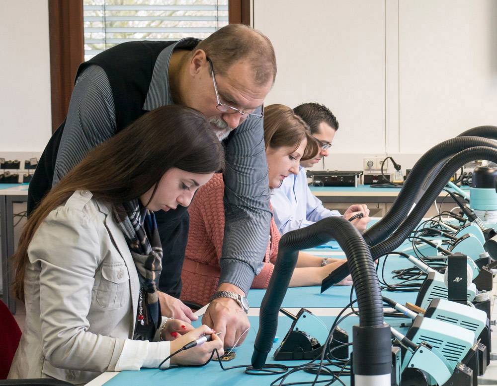 Een leraar helpt een studente bij een soldeerles in een elektronicalachtergrond, terwijl andere studenten geconcentreerd aan hun projecten werken.