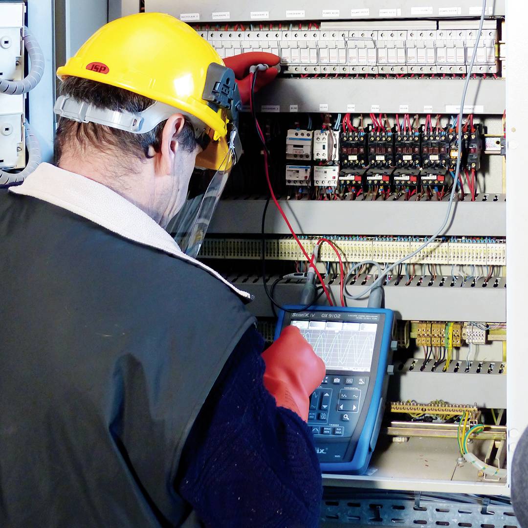 Een technicus met beschermhelm en -schild werkt aan een elektrisch schakelbord met een oscilloscoop-achtig apparaat.