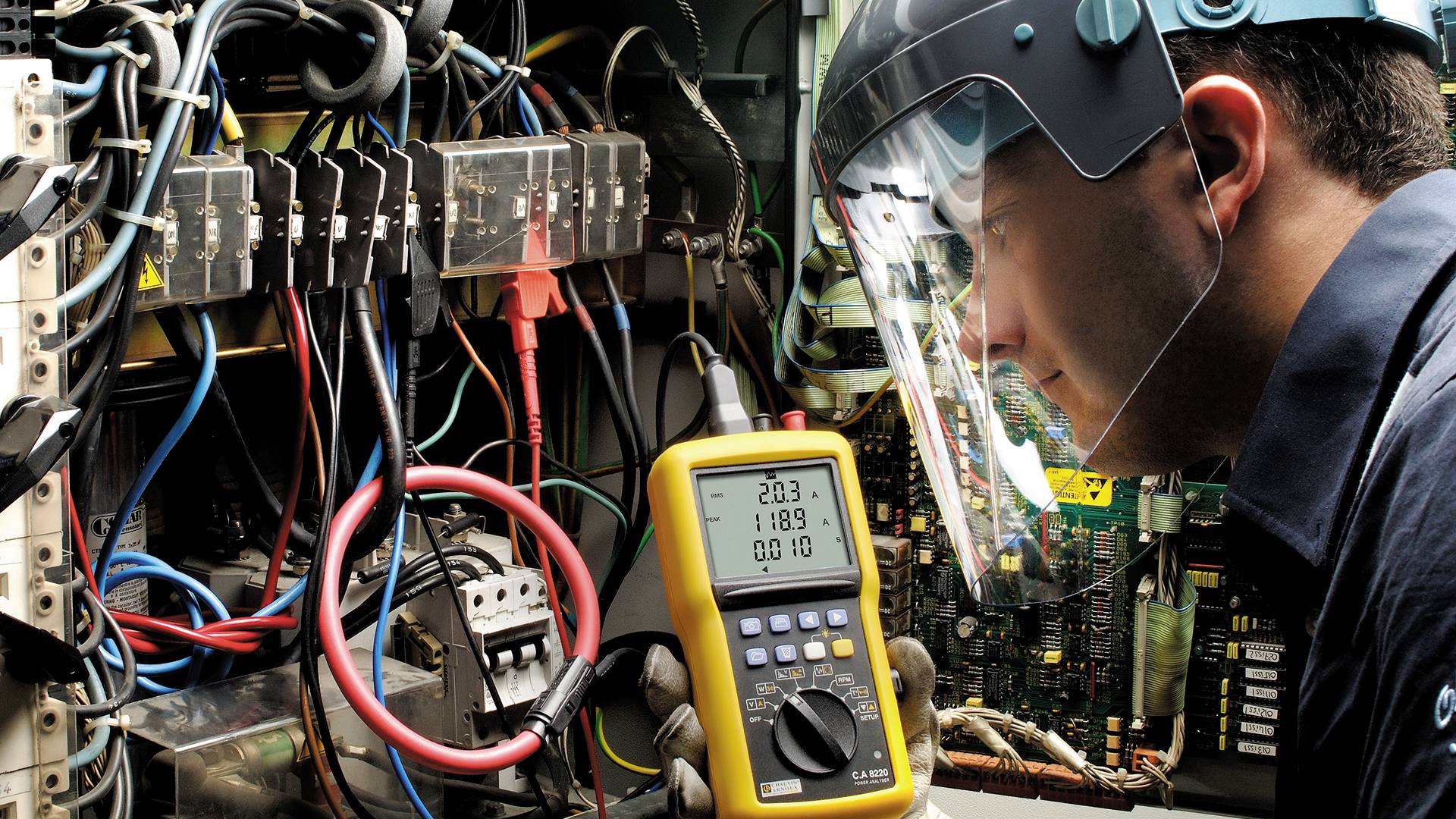 Een technicus meet de elektrische stroom in een verdeelkast met een digitaal multimeter. Hij draagt een beschermingsmasker.