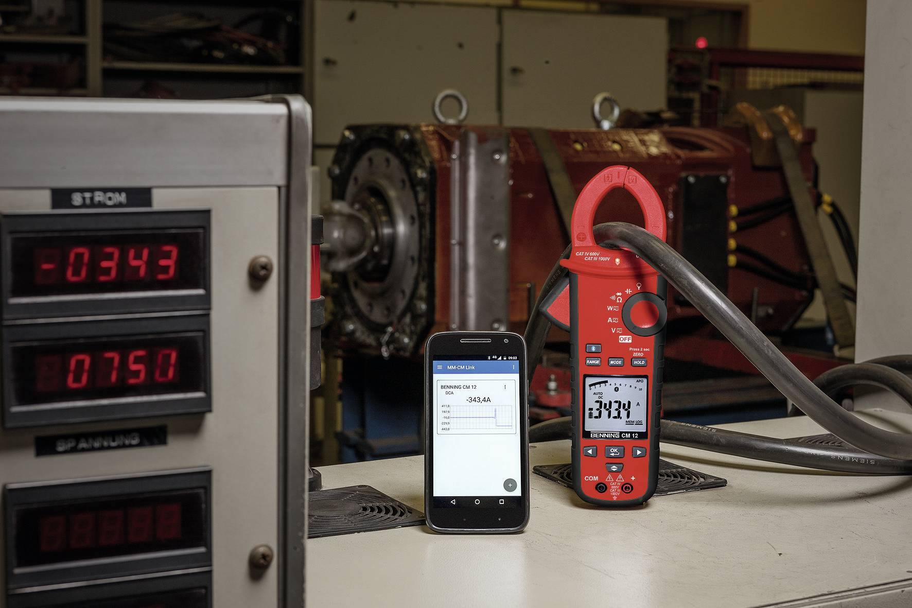 Een digitale multimeter meet de stroom in een werkplaats. Een smartphone toont dezelfde meting. Op de achtergrond een machine.
