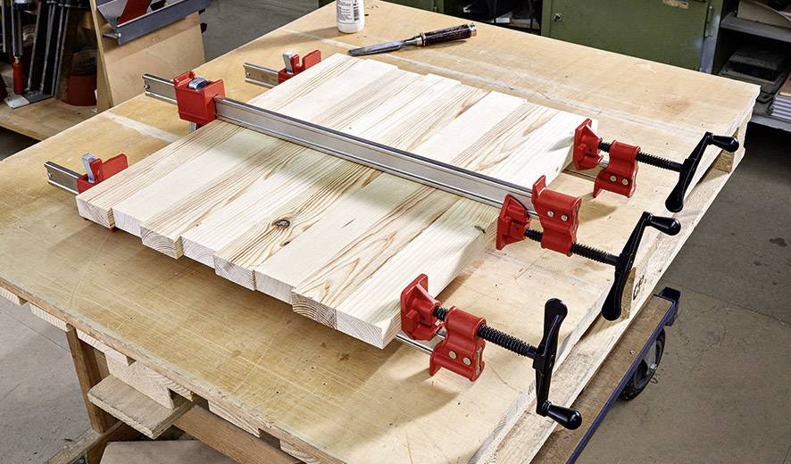 Houten stukken worden met vier rode klemmen op een werkbank vastgezet om verlijmd te worden.