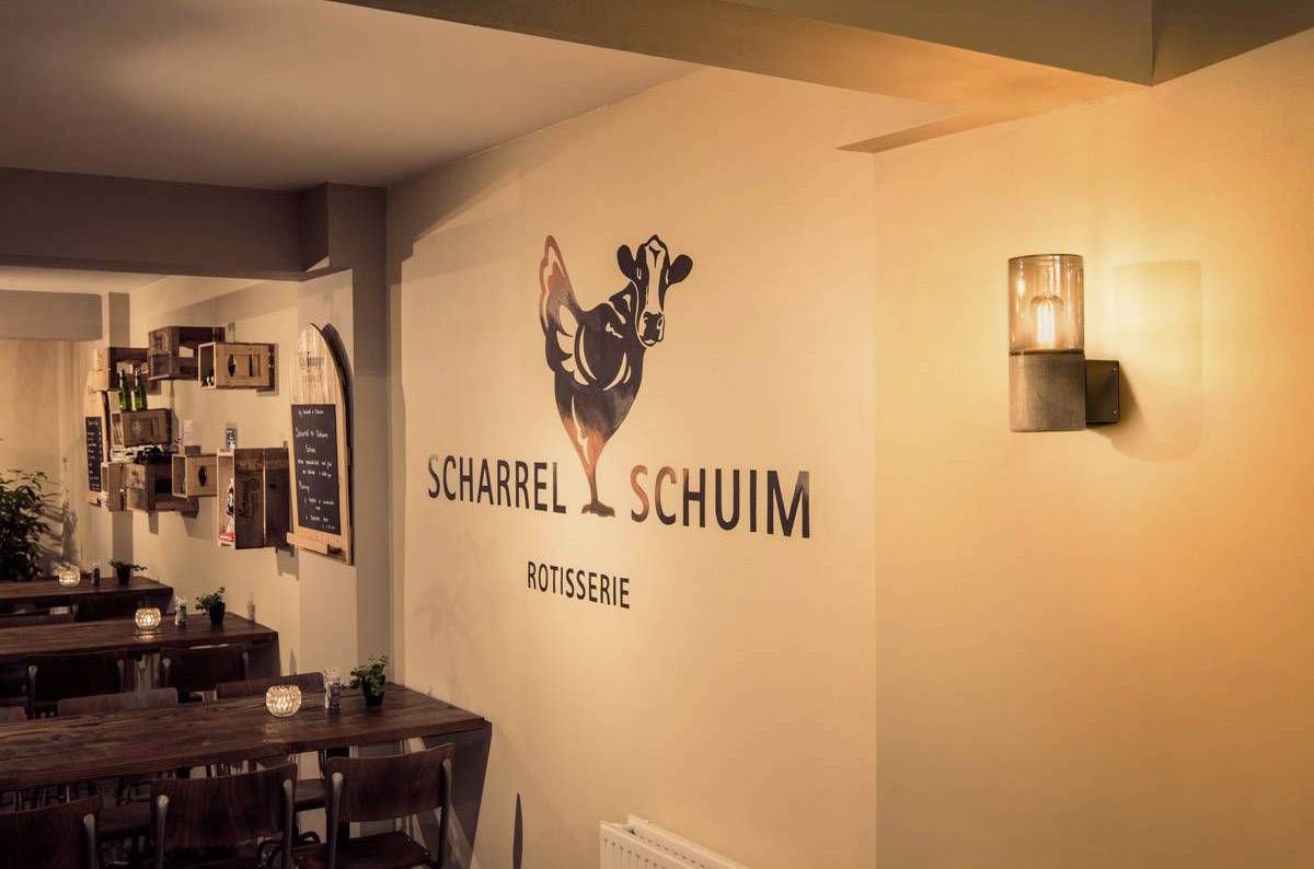 Een restaurant met de tekst 'Scharrel Schuim Rotisserie' aan de muur, houten tafels en gedempte verlichting creëren een gezellige sfeer.
