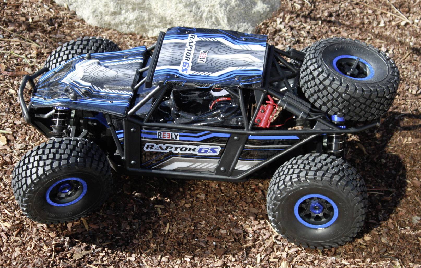 Een RC-buggy met het opschrift 'Raptor 6S' en 'Arrma' in blauw en zwart, staat op de grond. De auto heeft grote, grove banden.